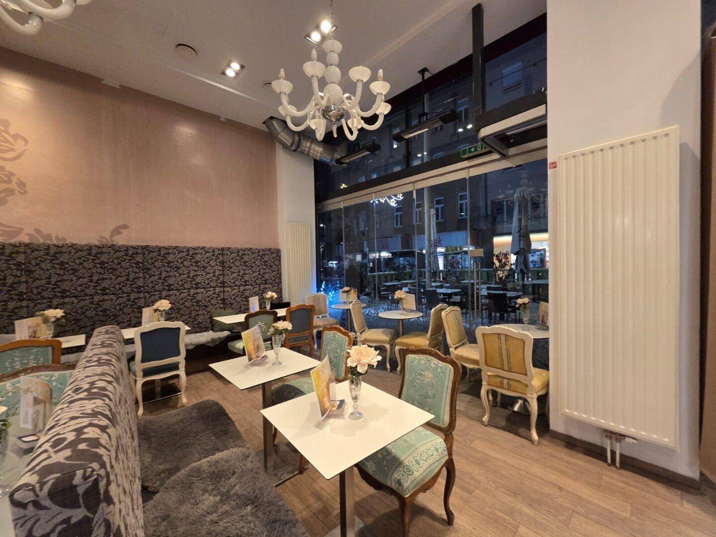 Gemütlicher Café-Innenraum mit eleganten Sitzmöbeln, Kronleuchtern und einem großen Heizkörper unter den Fenstern.