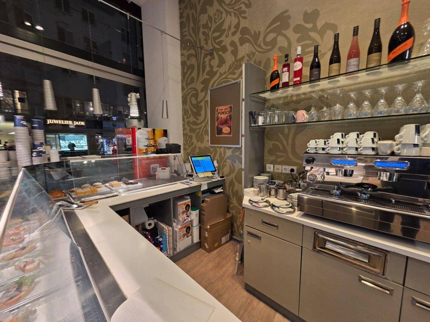 Thekenbereich eines Cafés mit Kaffeemaschine, Vitrine und Regalen für Gläser und Flaschen.