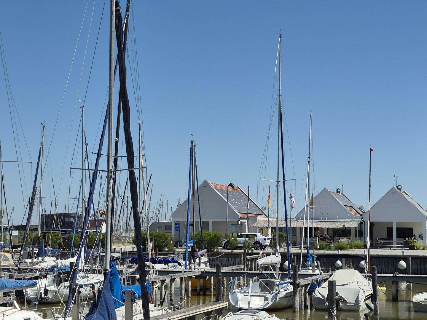 Sonniger Yachthafen mit Segelbooten an Stegen und modernen Gebäuden mit Solaranlagen im Hintergrund.