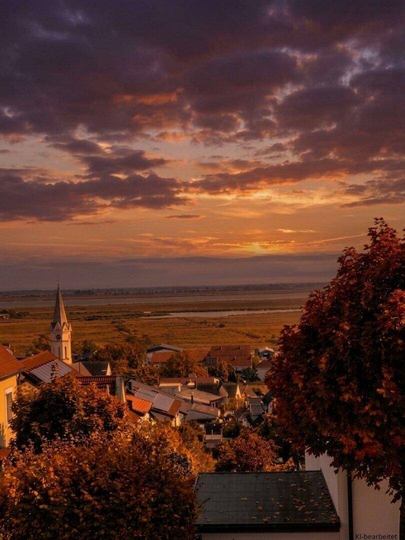 Malerischer Ausblick auf ein Dorf mit Kirche, Herbstbäumen und einem See bei Sonnenuntergang.