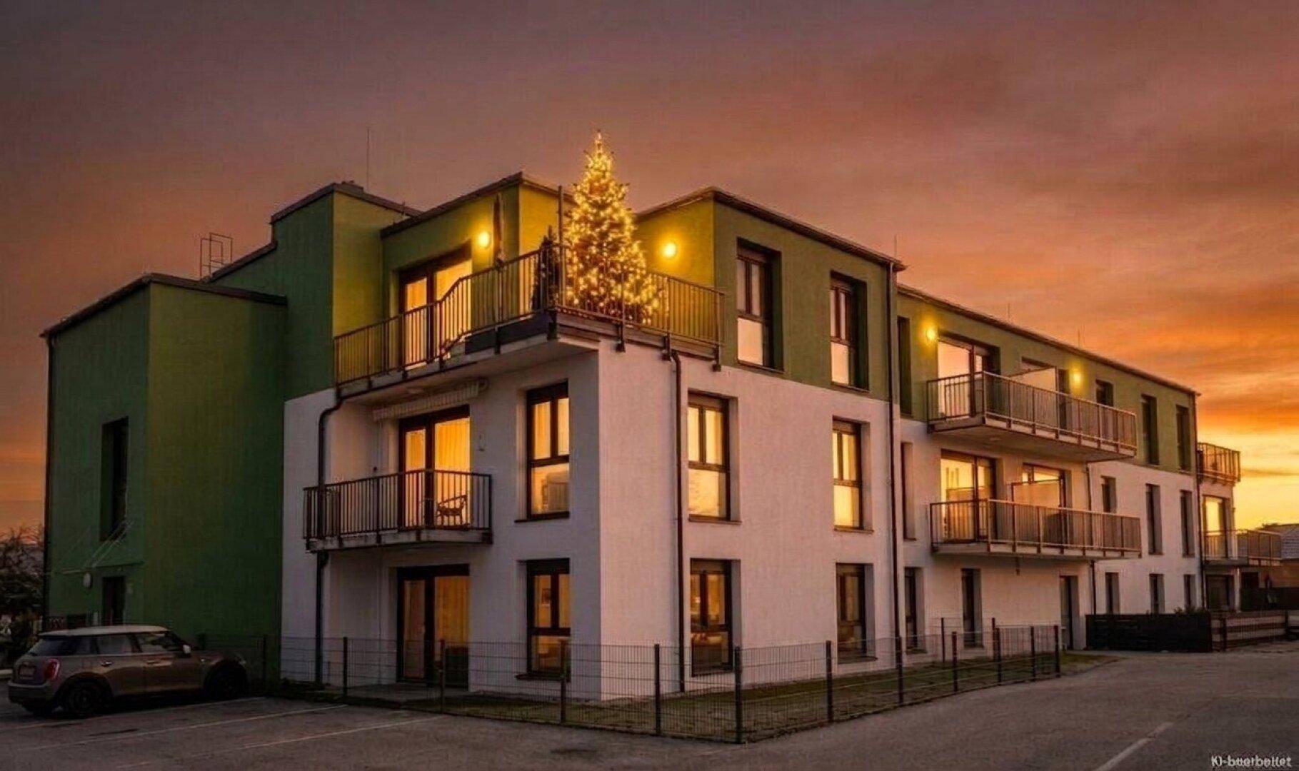 Modernes Mehrfamilienhaus mit Balkonen und einem festlich beleuchteten Weihnachtsbaum auf der Dachterrasse bei Sonnenuntergang.