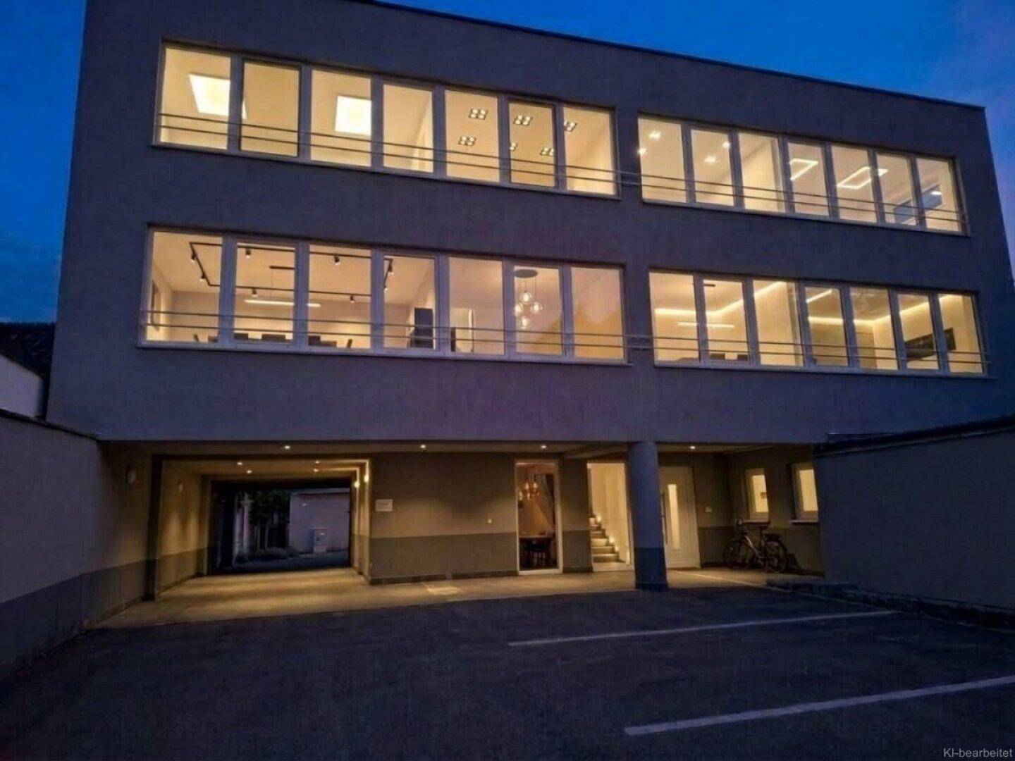 Moderne Gebäudefassade bei Nacht mit großen, beleuchteten Fenstern und Parkmöglichkeiten im Freien.