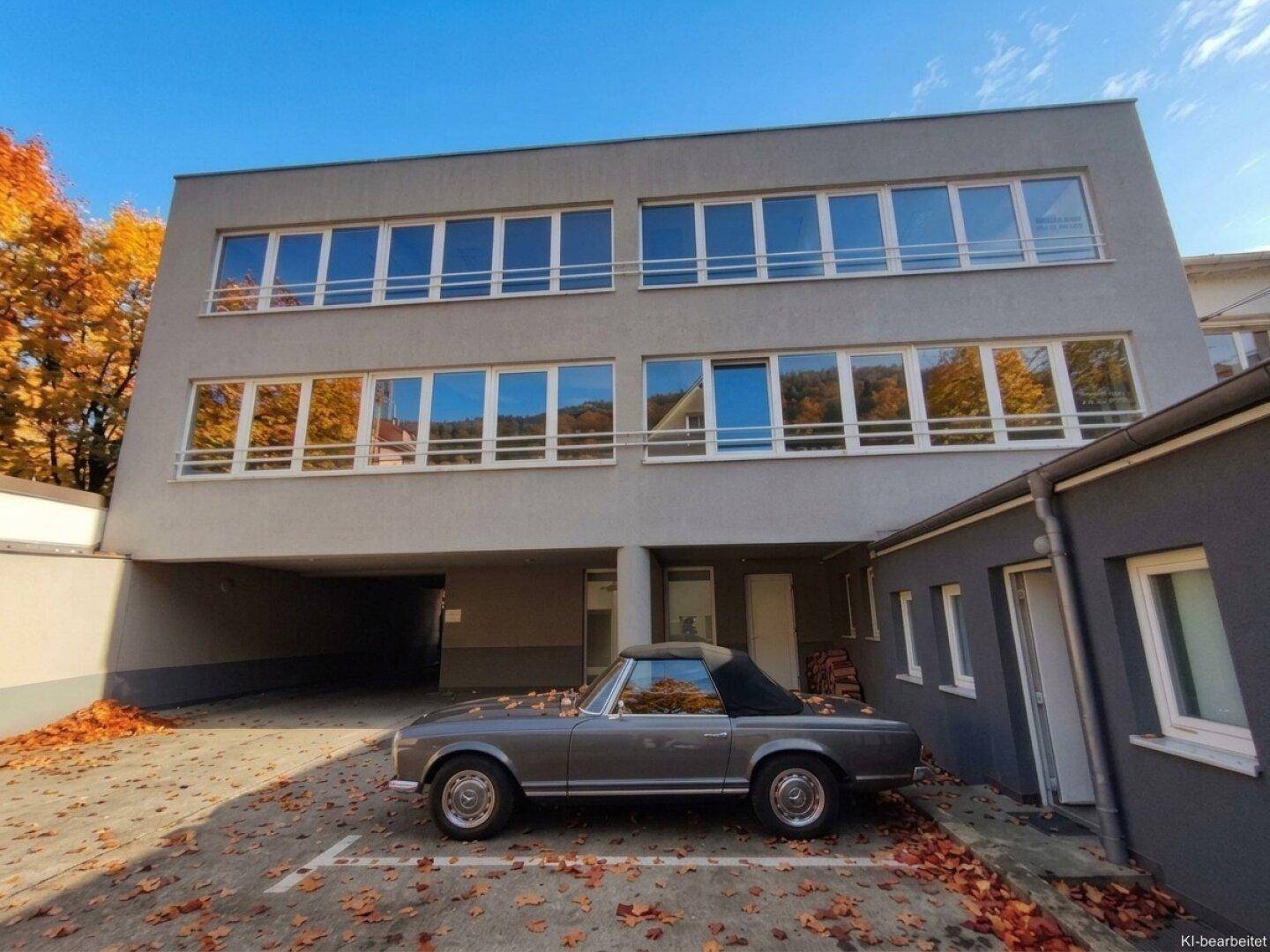 Modernes Gebäude mit großen Fenstern, überdachtem Parkplatz und Herbstlaub auf dem Boden.