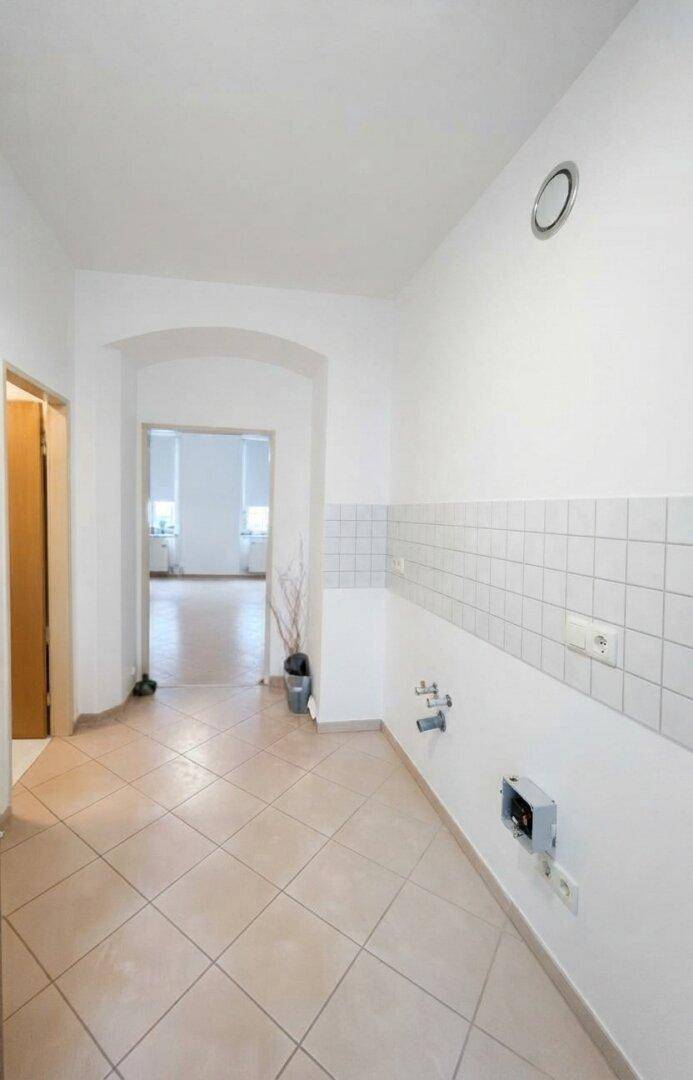 Leerer Raum mit Fliesenboden und vorbereiteten Anschlüssen für eine Küche, in gutem Zustand.