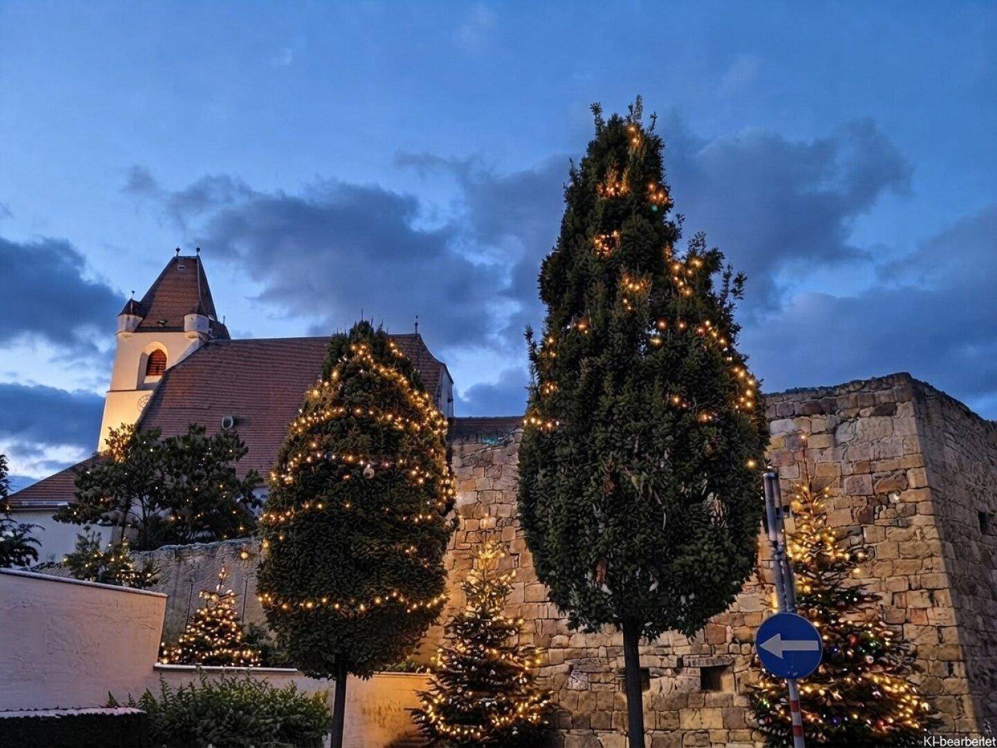 Abendliche Ansicht eines Gebäudes und einer Steinmauer mit festlich beleuchteten Weihnachtsbäumen.