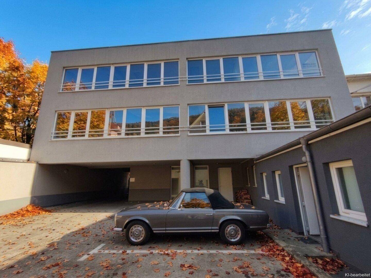 Modernes Gebäude mit großen Fenstern und einem Parkplatz, umgeben von herbstlichem Laub.