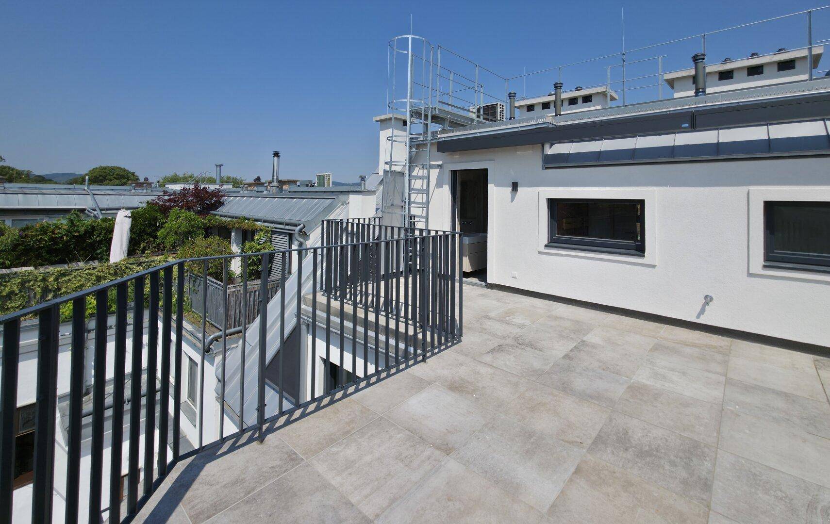 Eine weitläufige Dachterrasse mit Fliesenboden und Metallgeländer unter blauem Himmel.