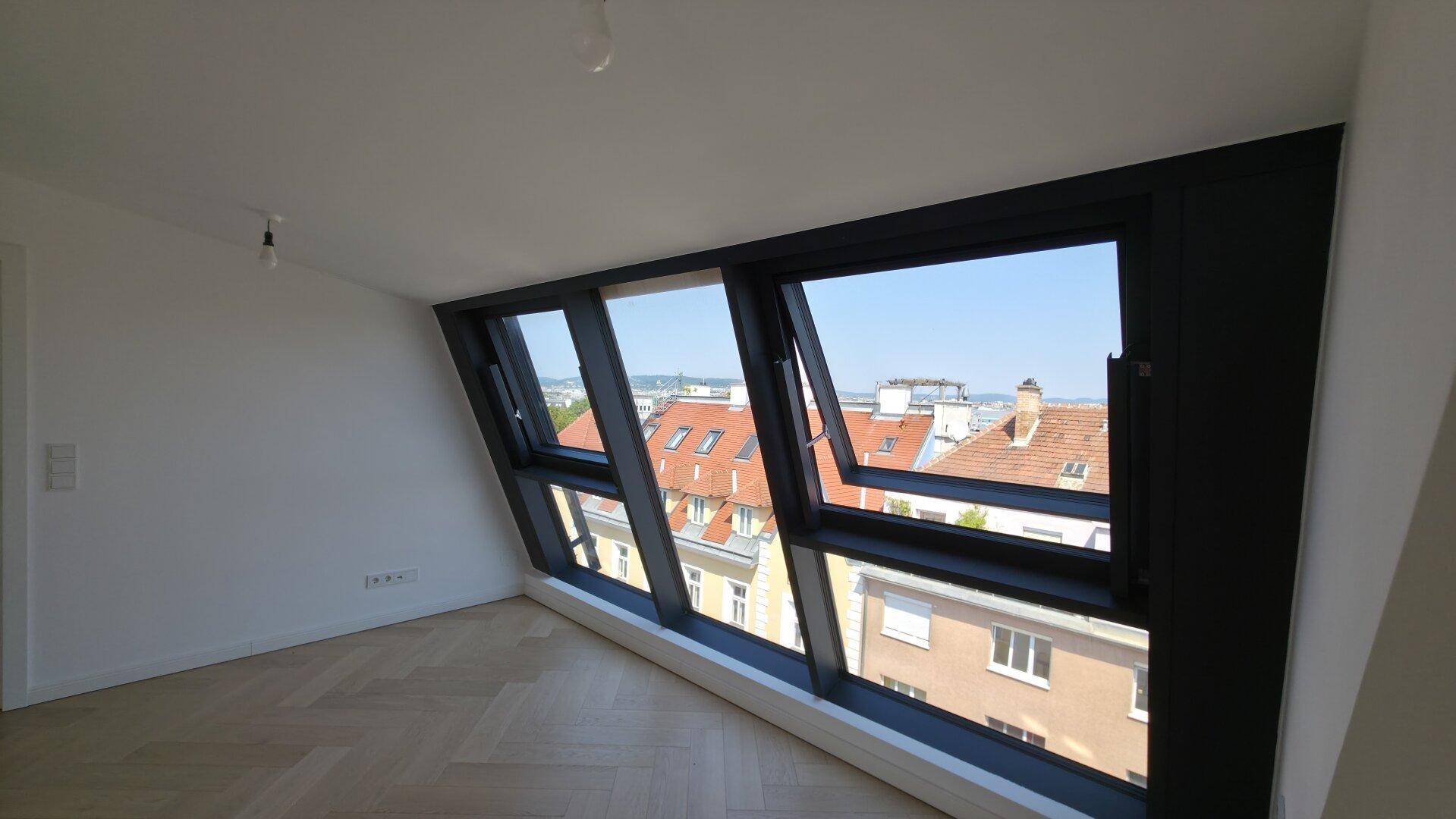 Heller Raum mit schrägen Wänden, großen Fenstern und Blick über die Dächer der Stadt.