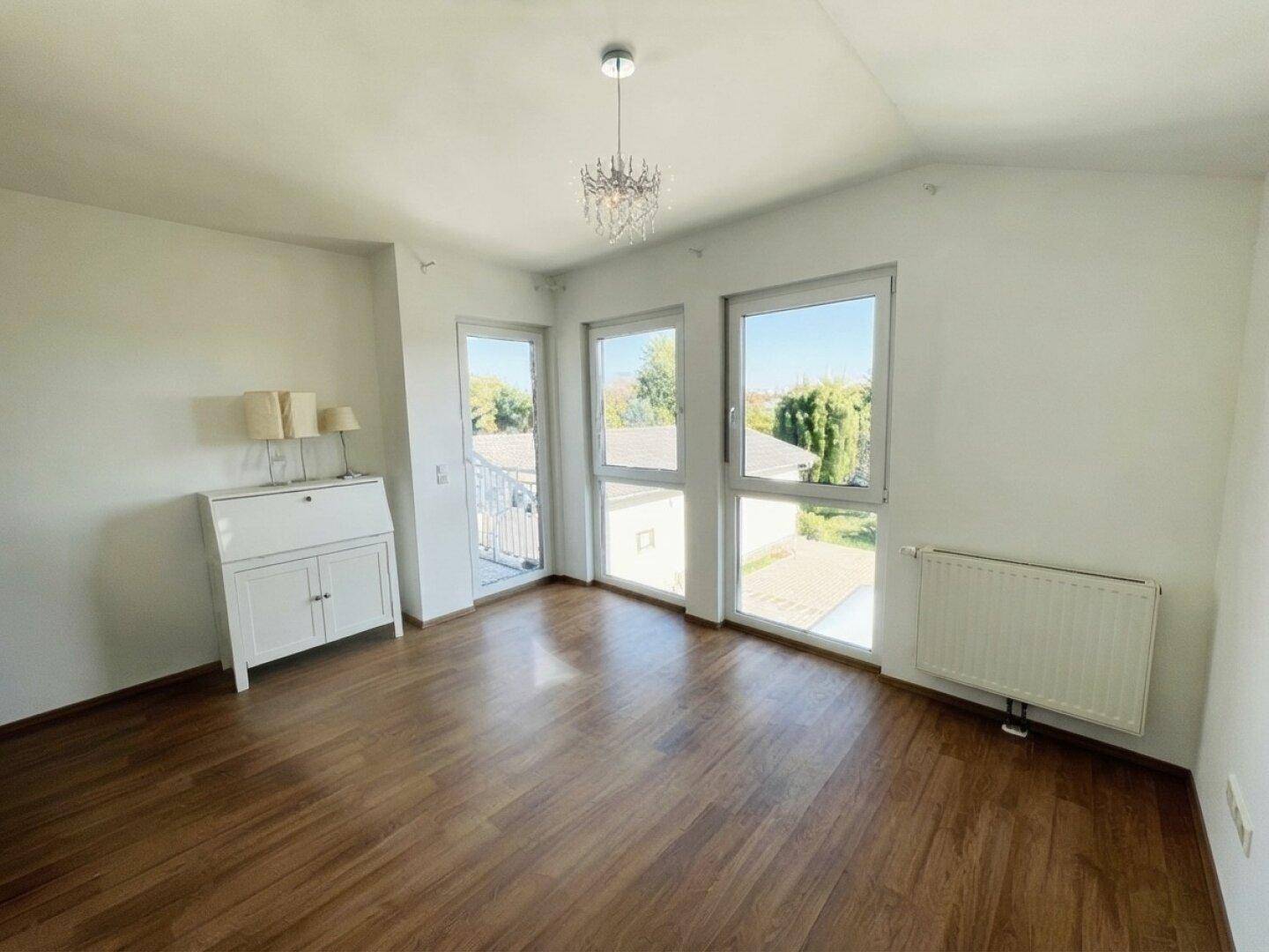 Helles Zimmer mit Holzboden, großen Fenstern und Zugang zu einem kleinen Balkon.