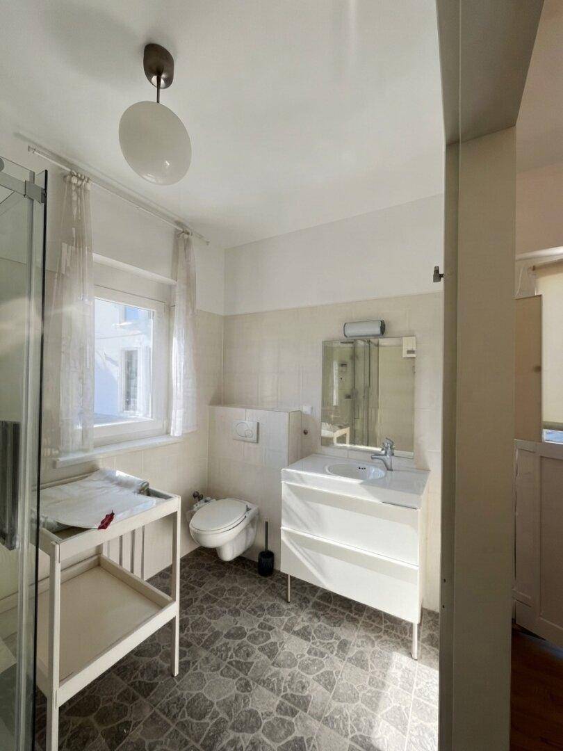 Badezimmer mit Toilette, Waschbecken und Duschbereich, hell gefliest und mit Fenster.