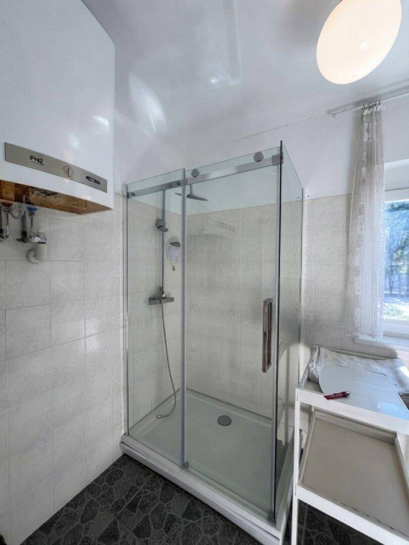 Helles Badezimmer mit moderner Duschkabine, weißen Wandfliesen, dunklem Boden und Fenster für Tageslicht.