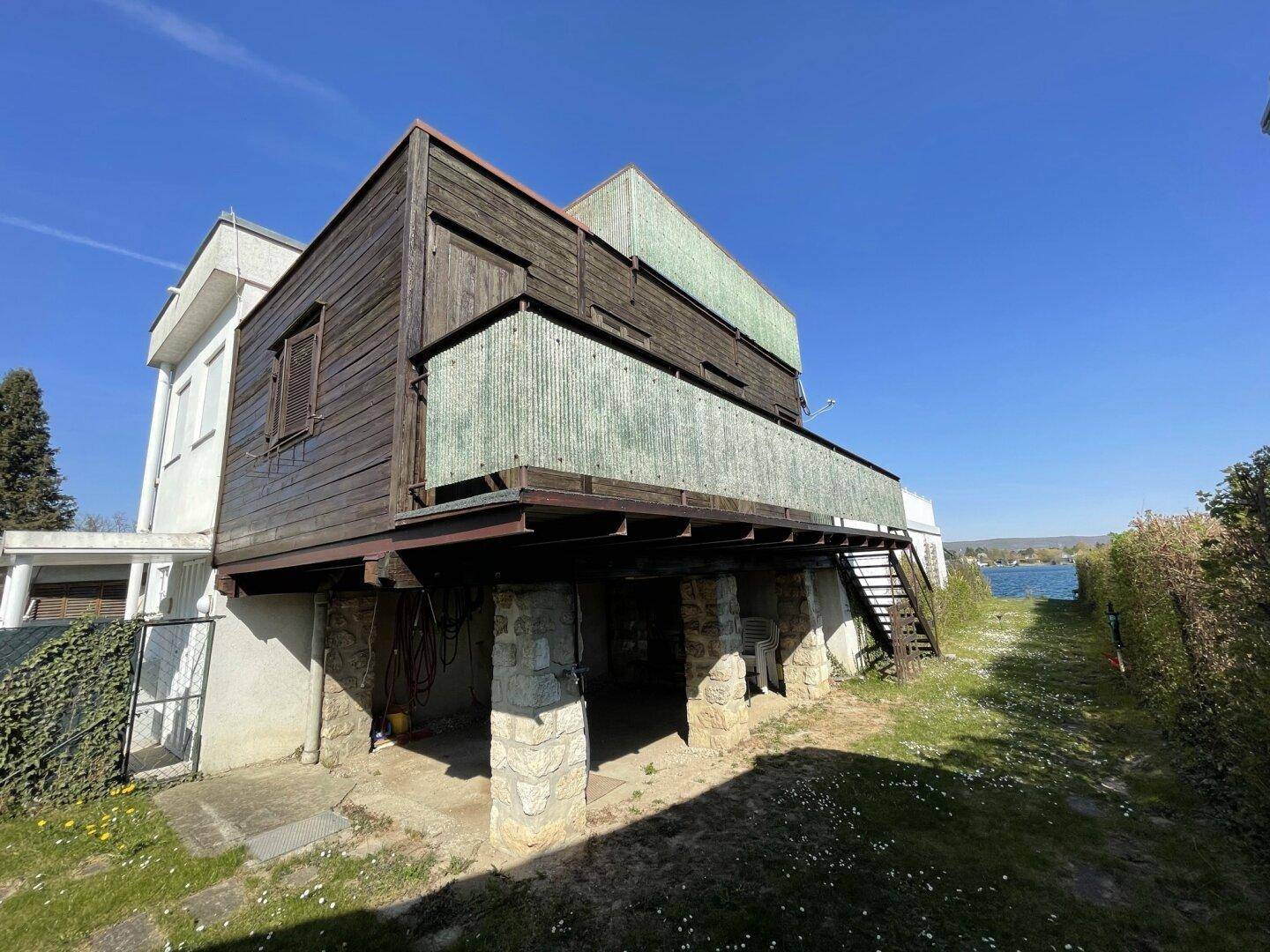 Gebäudeansicht mit erhöhter Holzkonstruktion auf Steinsäulen und Seeblick im Hintergrund.