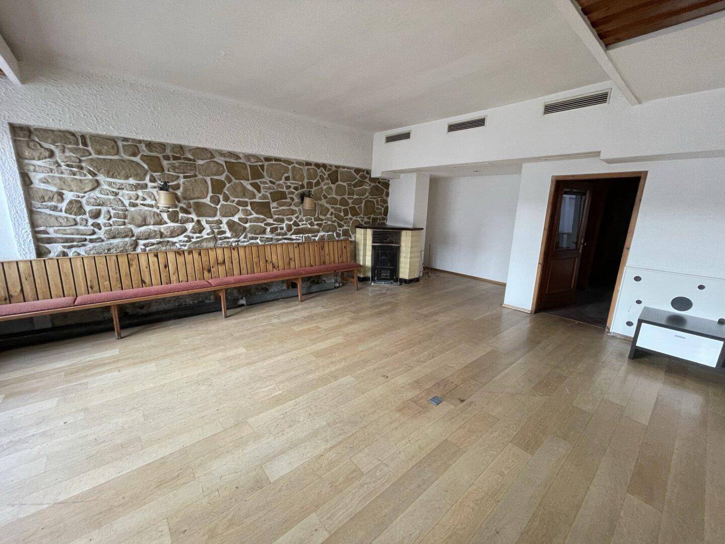 Geräumiger Raum mit hellem Holzfußboden, einer Steinwand und einem kleinen Ofen, ideal für gesellige Runden.