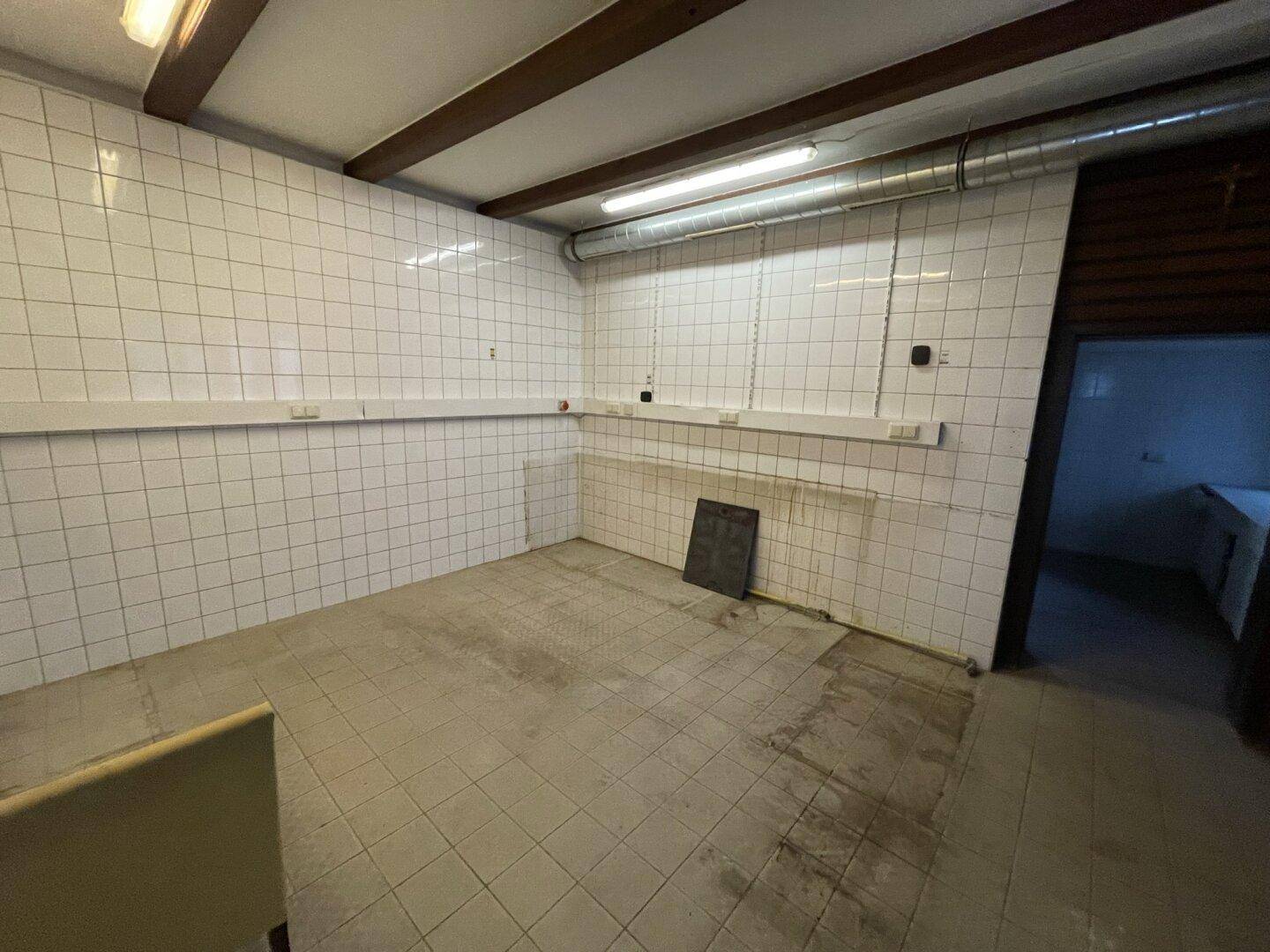 Funktionaler Raum mit weißen Fliesen an den Wänden, sichtbaren Deckenbalken und Lüftungsrohren.