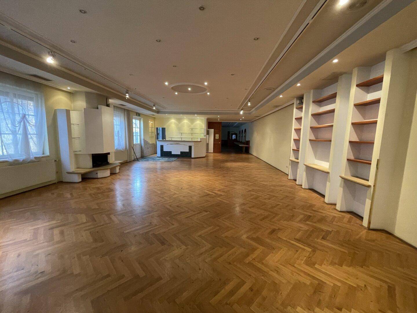 Geräumiger Saal mit Fischgrätparkett, integrierter Bar und dekorativem Kamin, ideal für Veranstaltungen.