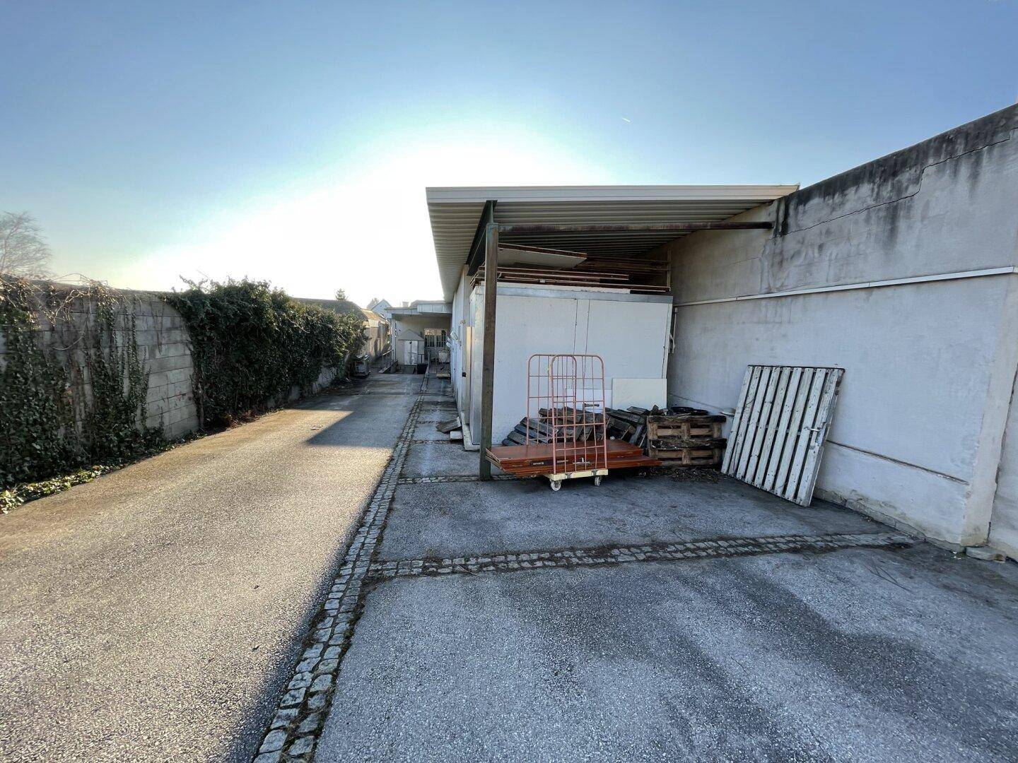 Außenbereich mit gepflastertem Weg, überdachtem Lagerbereich und einer Mauer mit Efeu.
