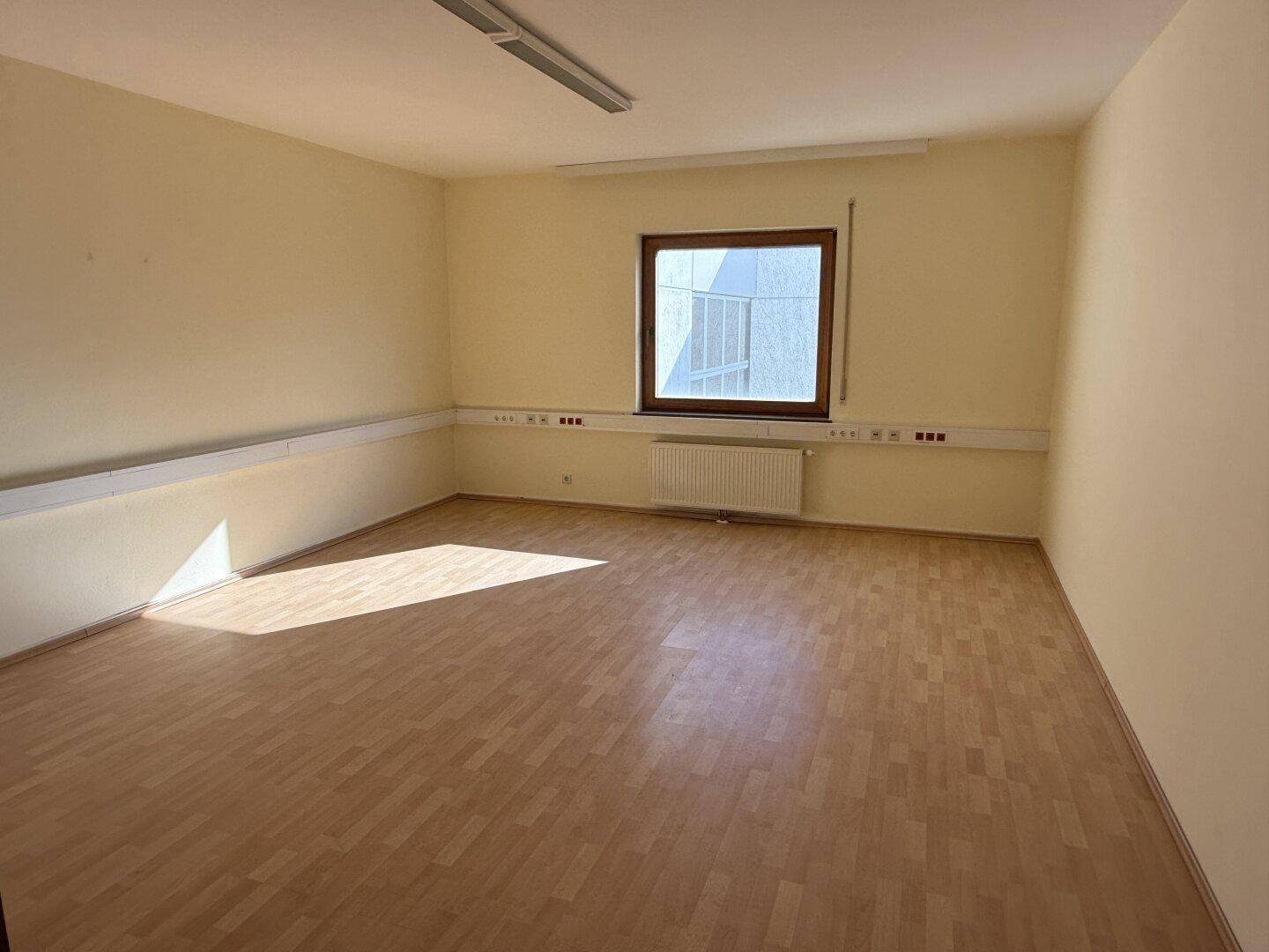 Kleinerer Büroraum mit Laminatboden, einem Fenster und Heizkörper, gut beleuchtet durch Tageslicht.