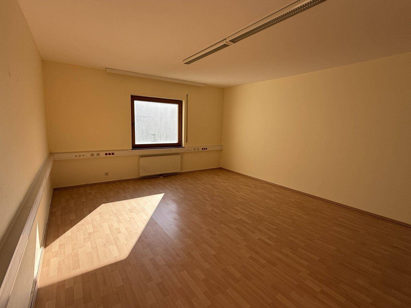 Leerer Büroraum mit hellem Laminatboden und einem Fenster, das viel Sonnenlicht hereinlässt.