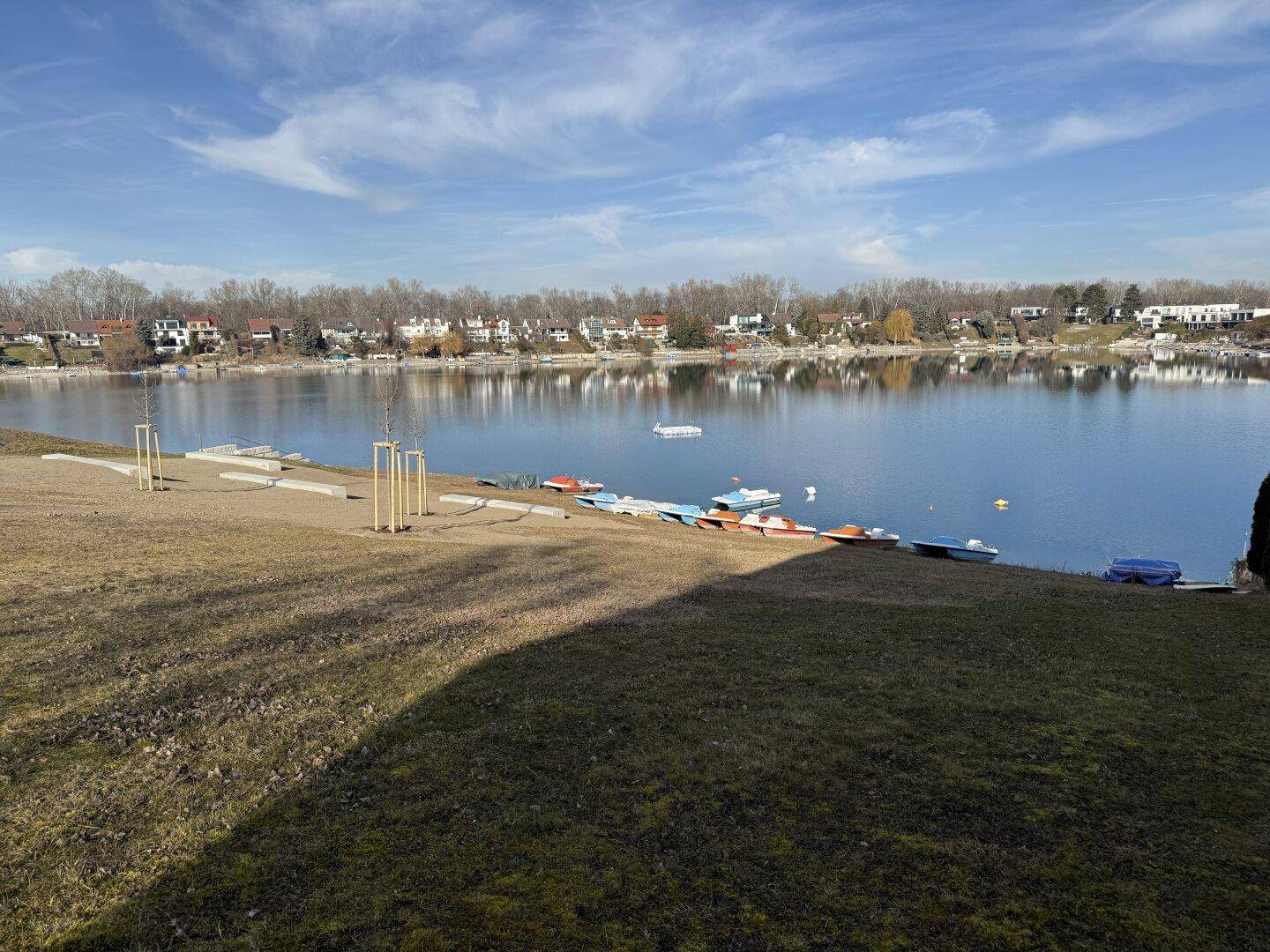 Panoramablick auf einen ruhigen See mit Booten am Ufer und Häusern in der Ferne.