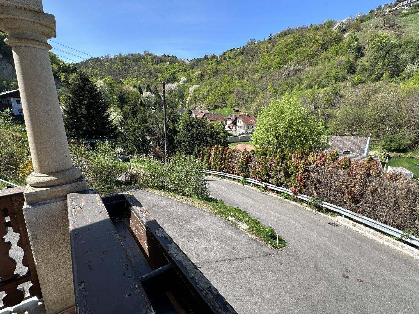 Blick vom Balkon auf die Straße und die umliegende grüne Hügellandschaft unter blauem Himmel.