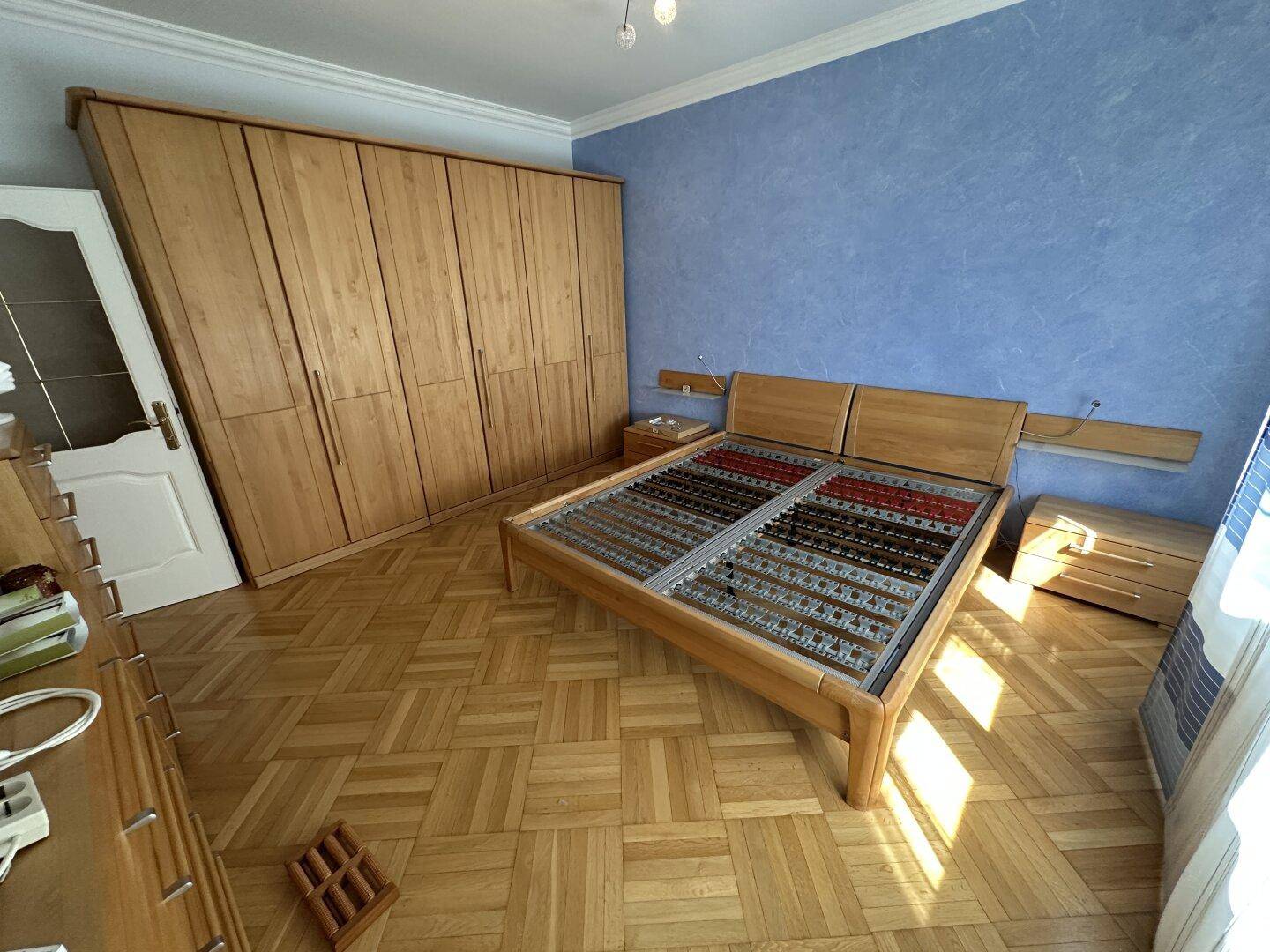 Schlafzimmer mit Fischgrätparkett, einem Bettgestell und einem großen Holzschrank.