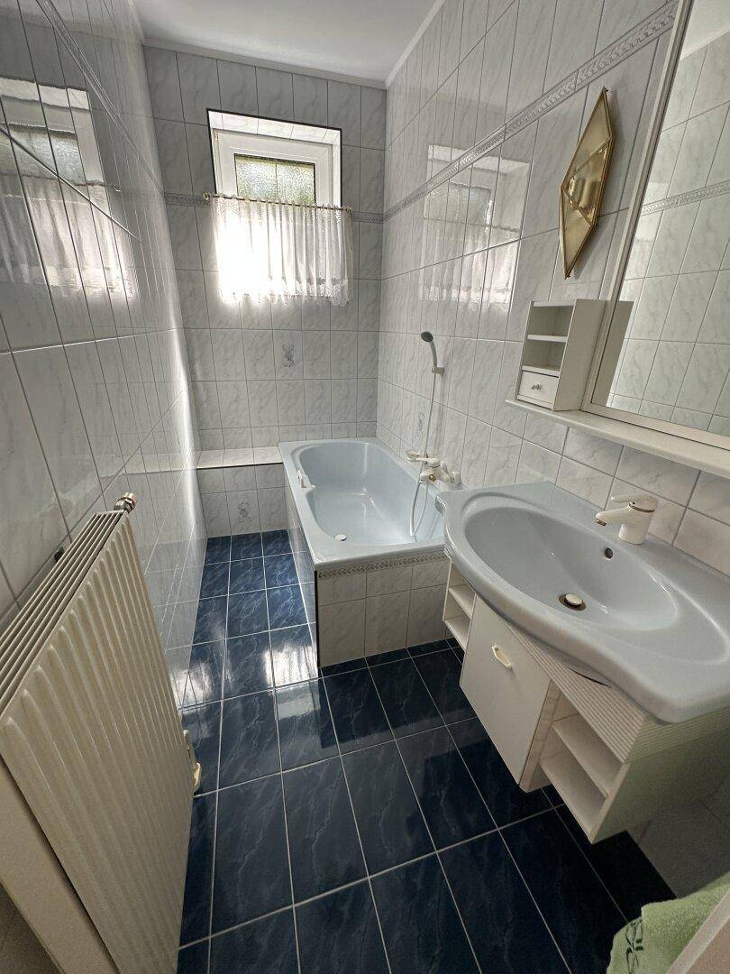 Gefliestes Badezimmer mit Badewanne, Waschbecken, Fenster und Heizkörper.