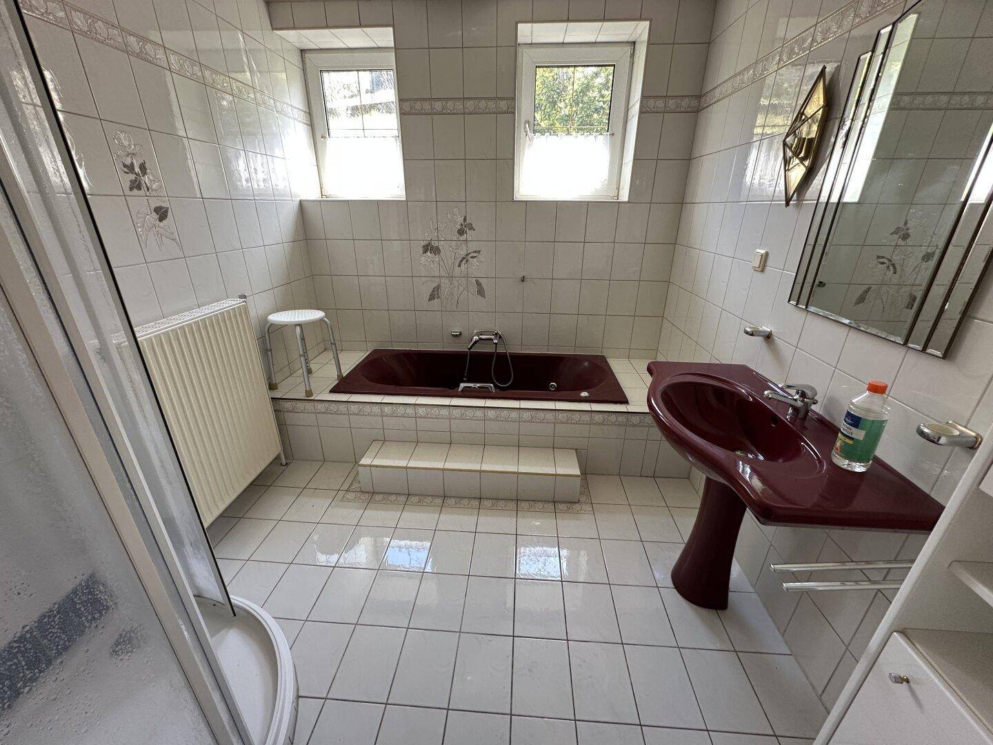 Badezimmer mit weißen Wandfliesen, einer roten Badewanne, Dusche und zwei Fenstern.