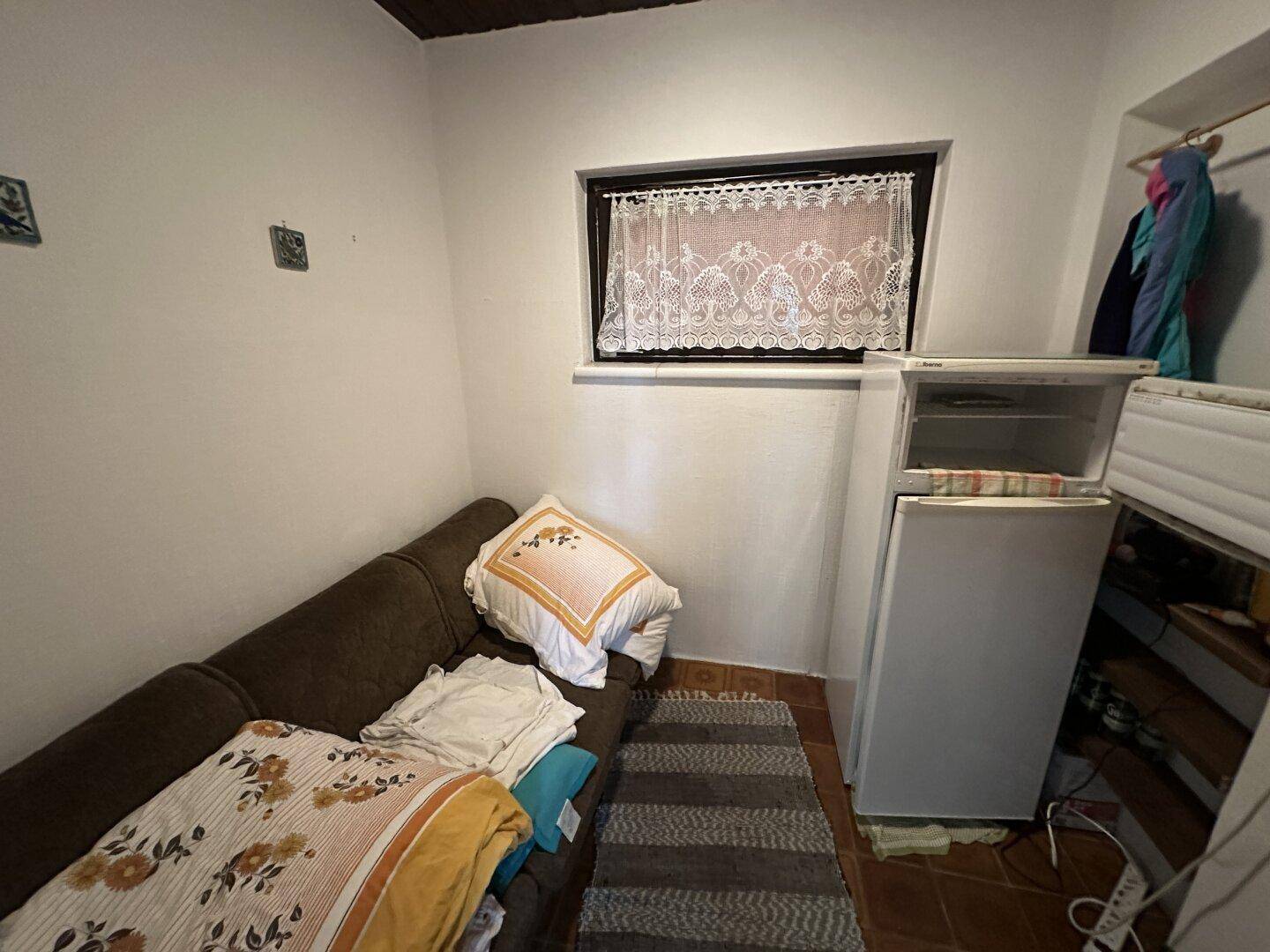 Kleiner Raum mit Sofa, Kühlschrank und einem Fenster mit Spitzenvorhang.