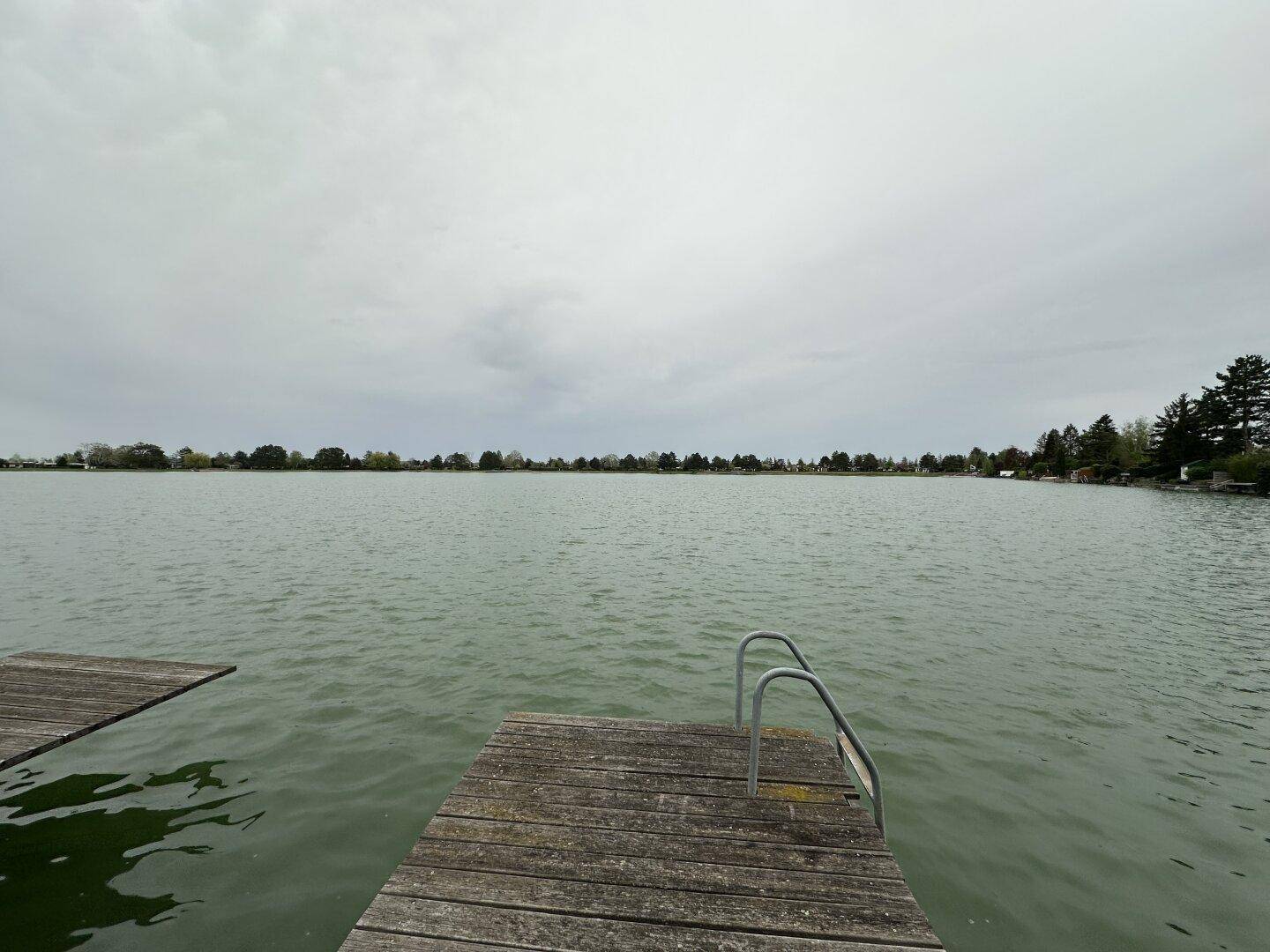 Weitläufiger Blick über einen ruhigen See mit einem Holzsteg im Vordergrund.