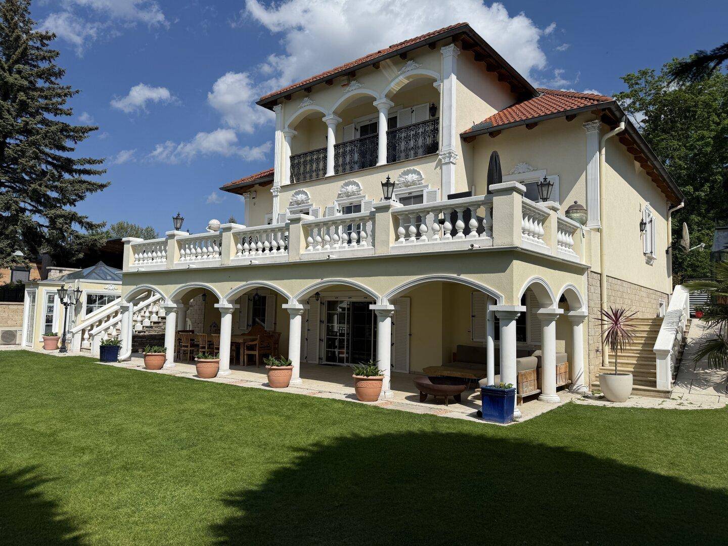 Prächtige Villa mit weitläufigem Garten, überdachter Terrasse und Balkonen unter blauem Himmel.