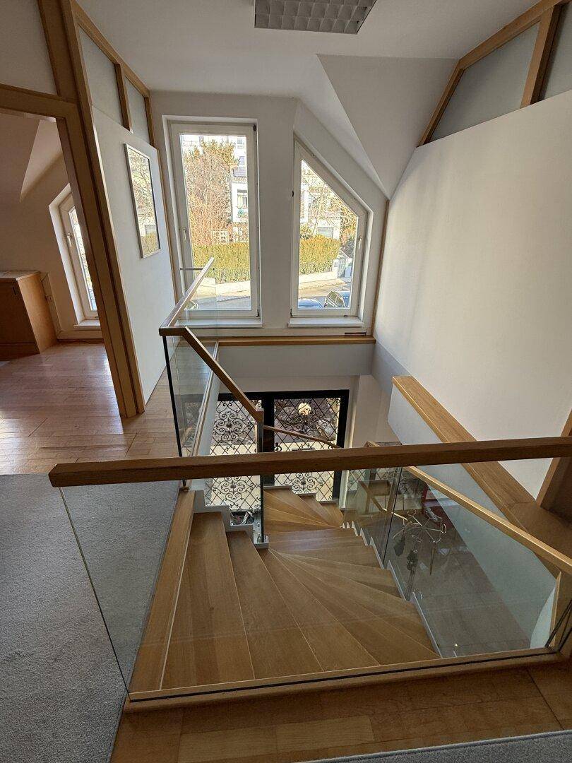 Elegante Holztreppe mit Glasgeländer, die zu den unteren Etagen führt, beleuchtet durch hohe Fenster.