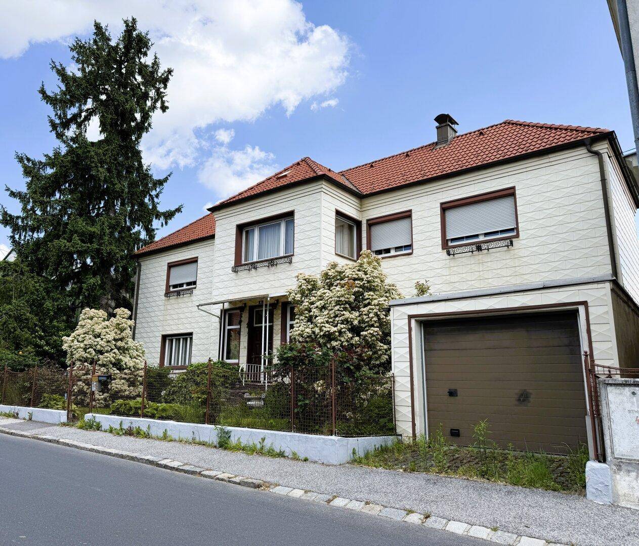 Einfamilienhaus mit integrierter Garage, rotem Ziegeldach und heller Fassade an der Straße.
