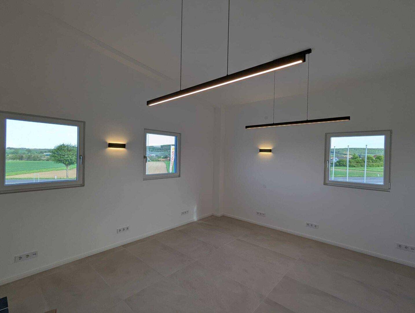 Heller, leerer Raum mit großen Fenstern, modernen Deckenleuchten und Fliesenboden, bietet weite Aussicht.