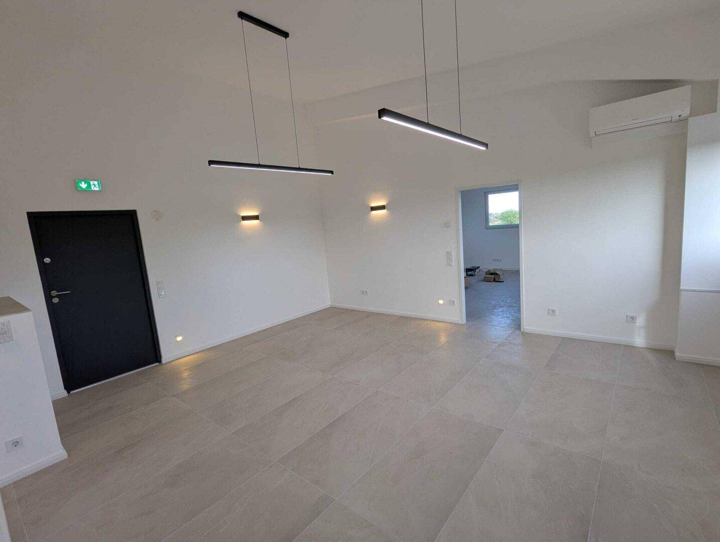 Leerer Raum mit hellem Fliesenboden, modernen Wand- und Deckenleuchten sowie einer dunklen Tür.