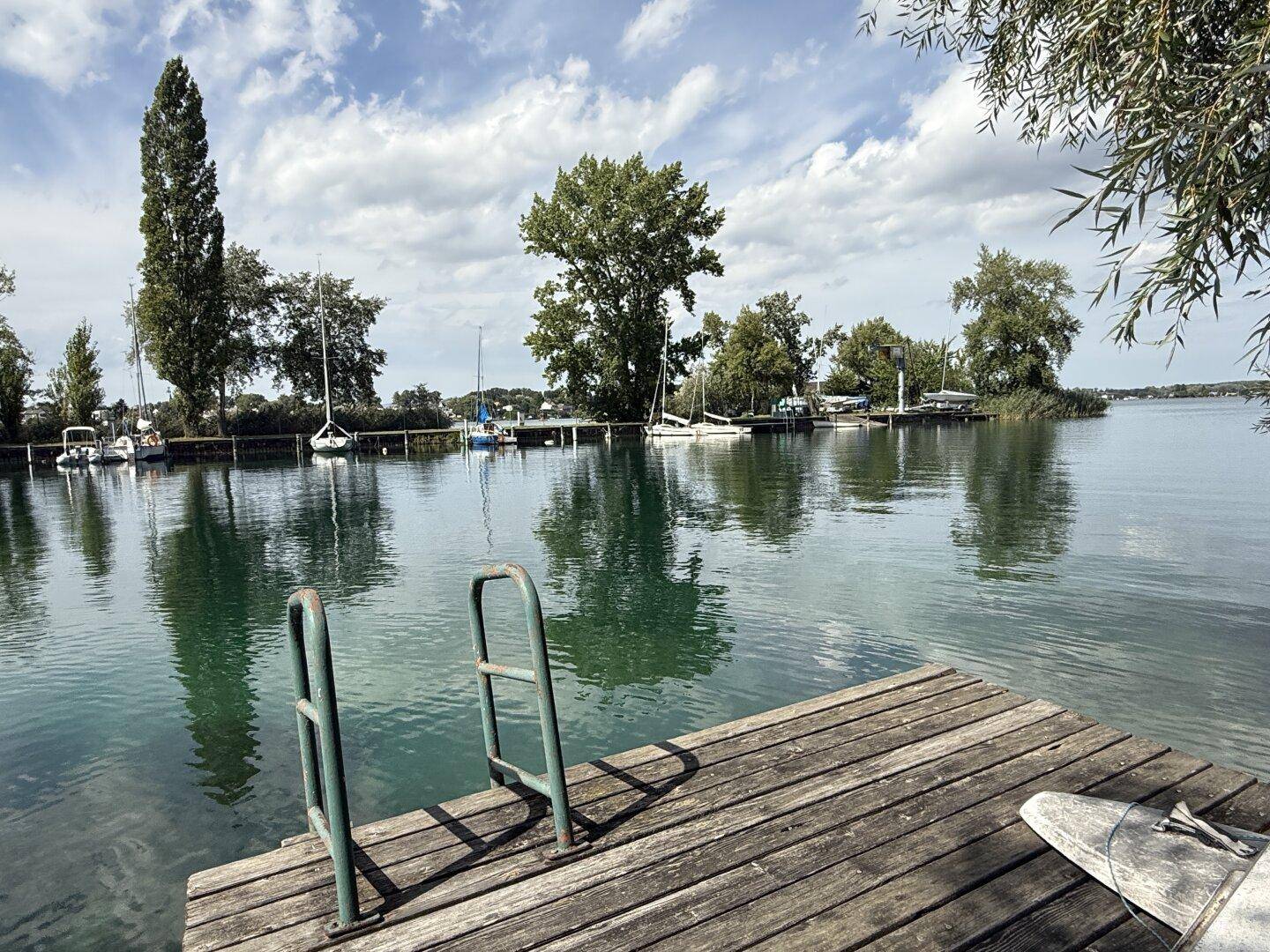 Weitläufiger Seeblick von einem Holzsteg mit Badeleiter und zahlreichen Segelbooten am Horizont.