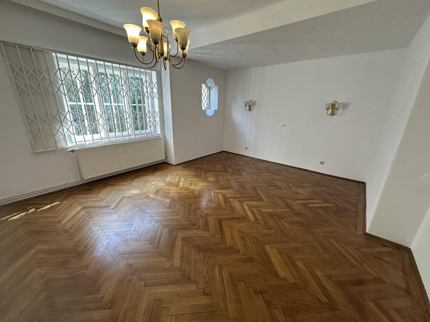 Helles Wohnzimmer mit elegantem Fischgrätparkett, weißen Wänden und einem großen Fenster mit Gitter.