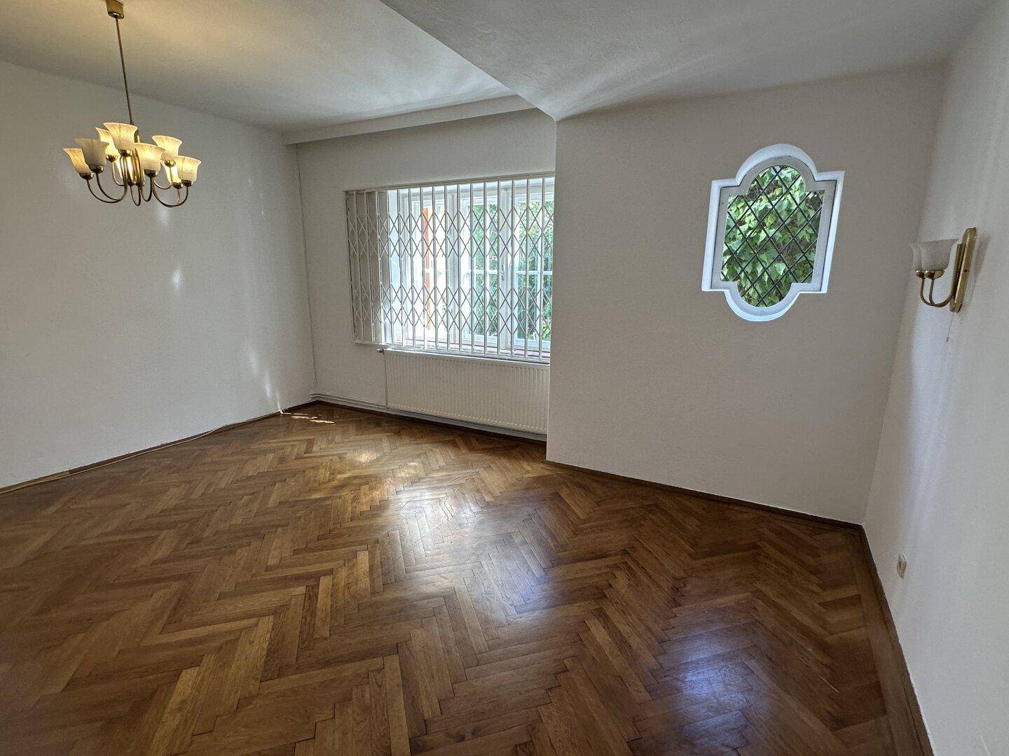 Wohnzimmer mit glänzendem Fischgrätparkett, weißen Wänden und einem Fenster mit Sicherheitsgitter.
