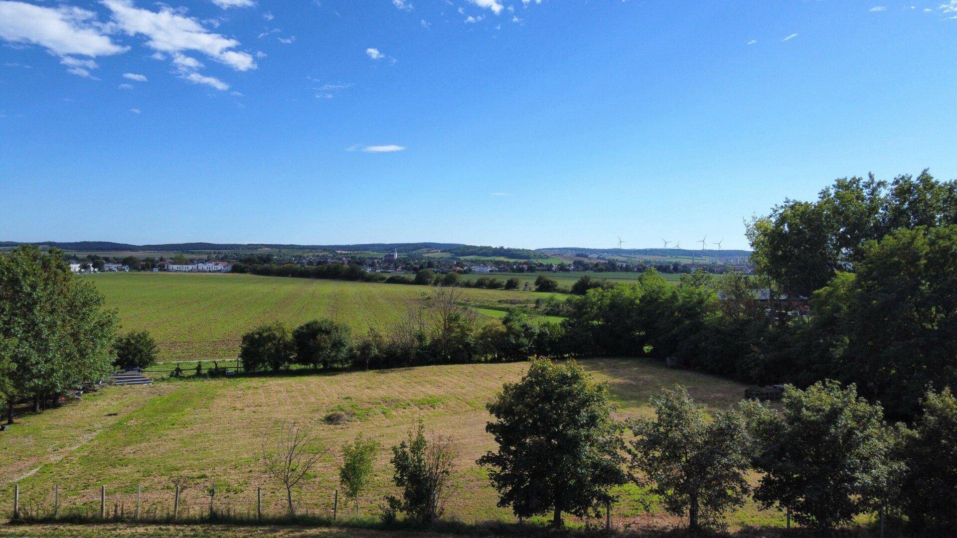 Weitläufige grüne Landschaft mit Feldern und einem Dorf in der Ferne unter blauem Himmel.