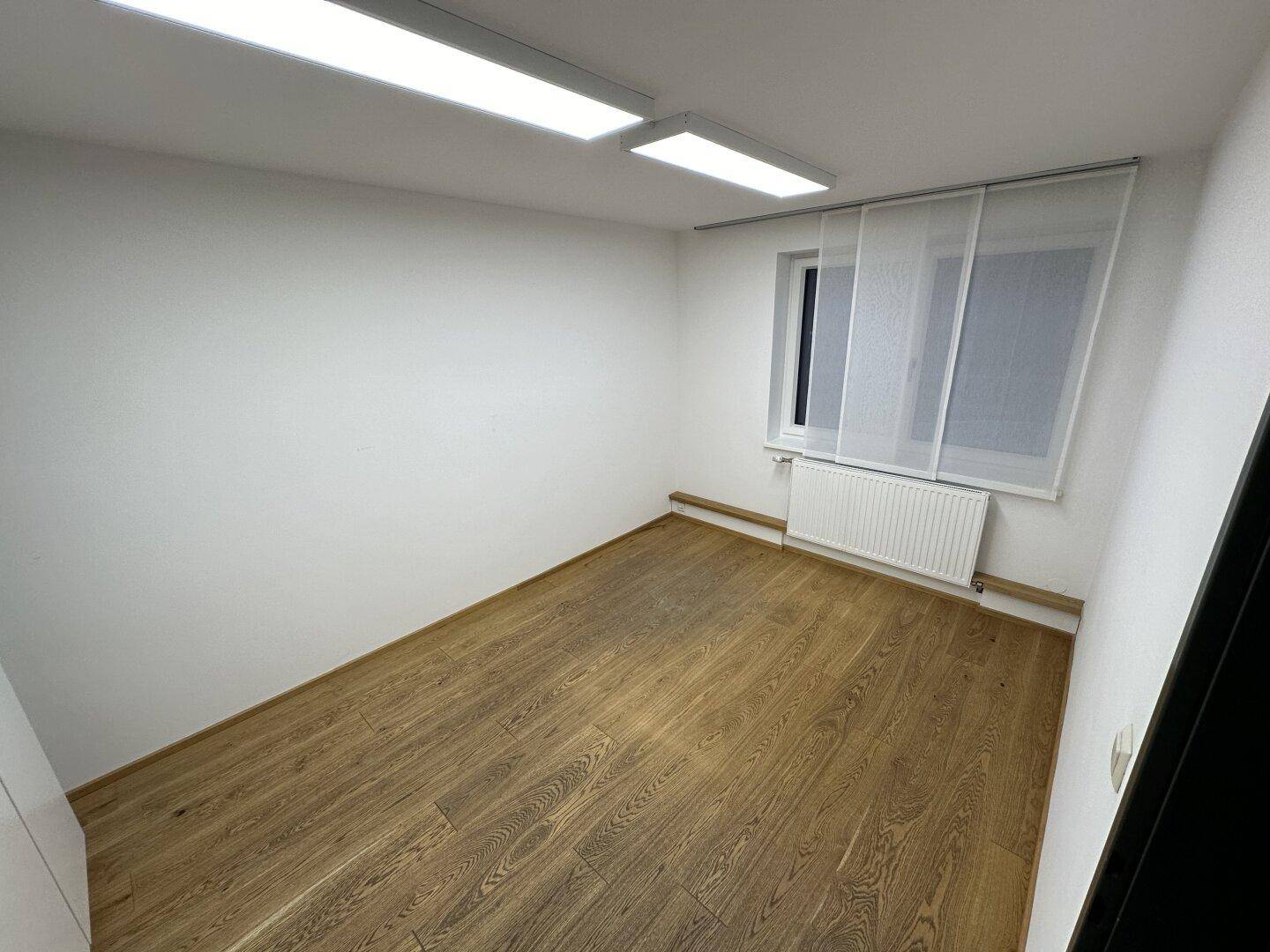 Helles, leeres Büro mit Holzboden, weißen Wänden und einem Fenster mit Heizkörper.
