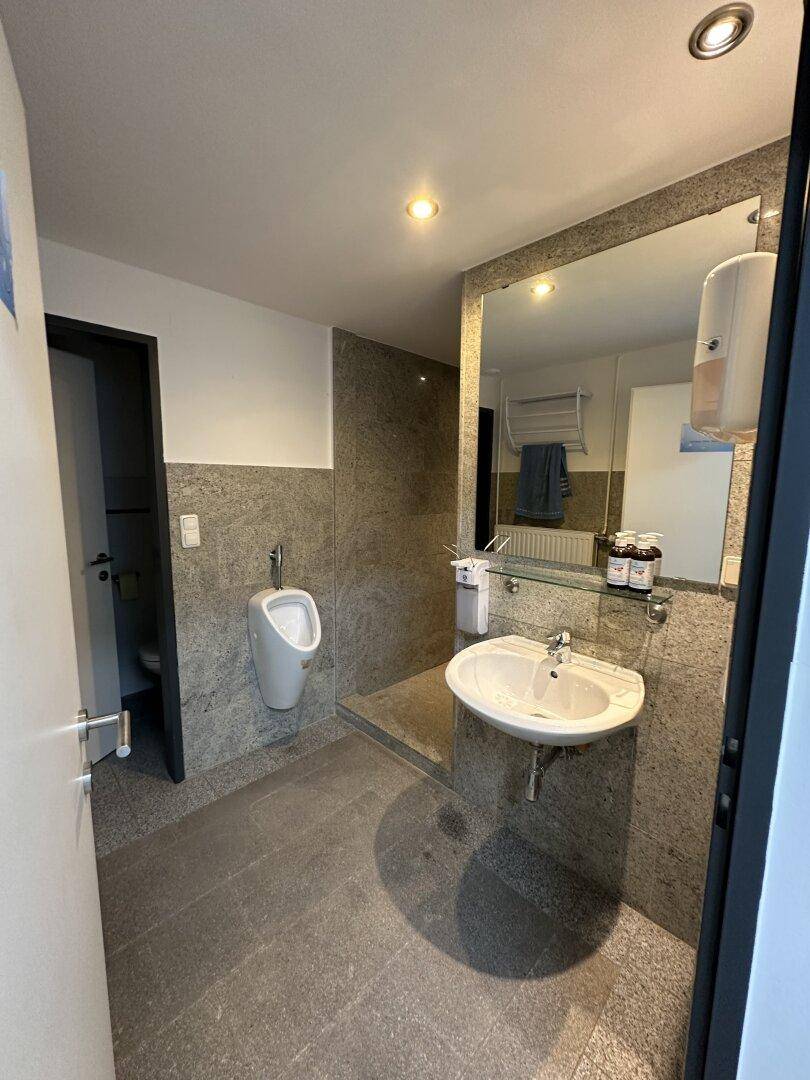 Modernes Badezimmer mit grauen Fliesen, weißem Waschbecken, großem Spiegel und Urinal.