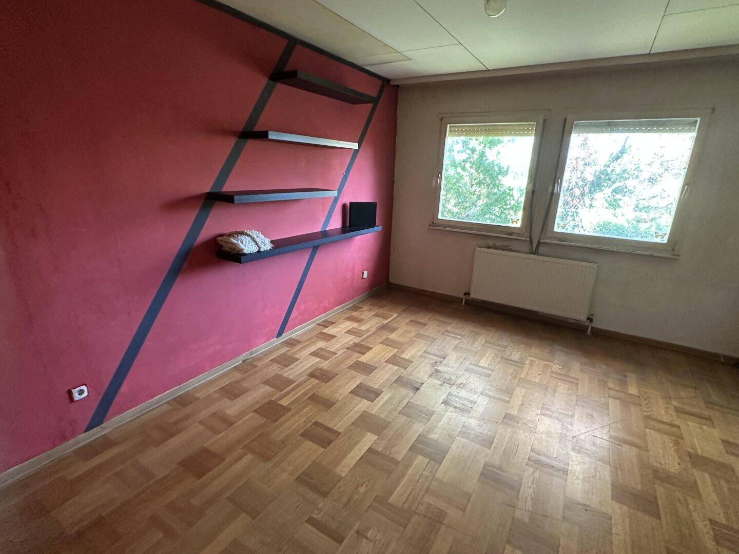 Schlafzimmer mit Parkettboden, roter Akzentwand mit Regalen und zwei Fenstern.