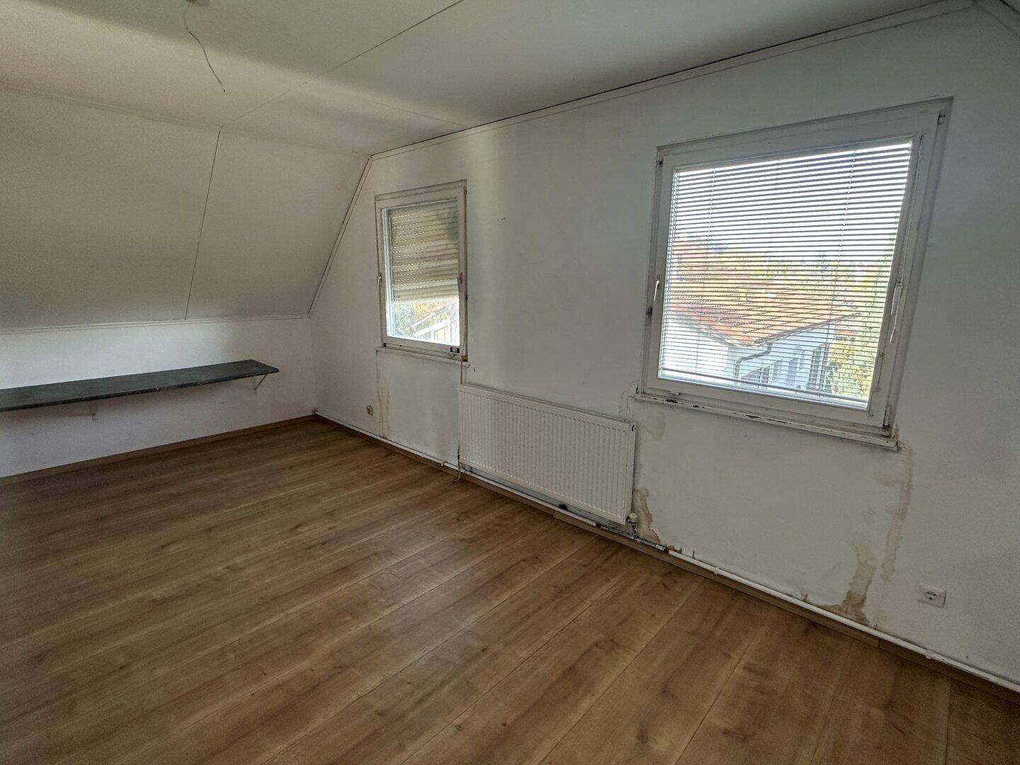 Dachzimmer mit Laminatboden, schrägen Wänden und zwei Fenstern, das renovierungsbedürftig ist.