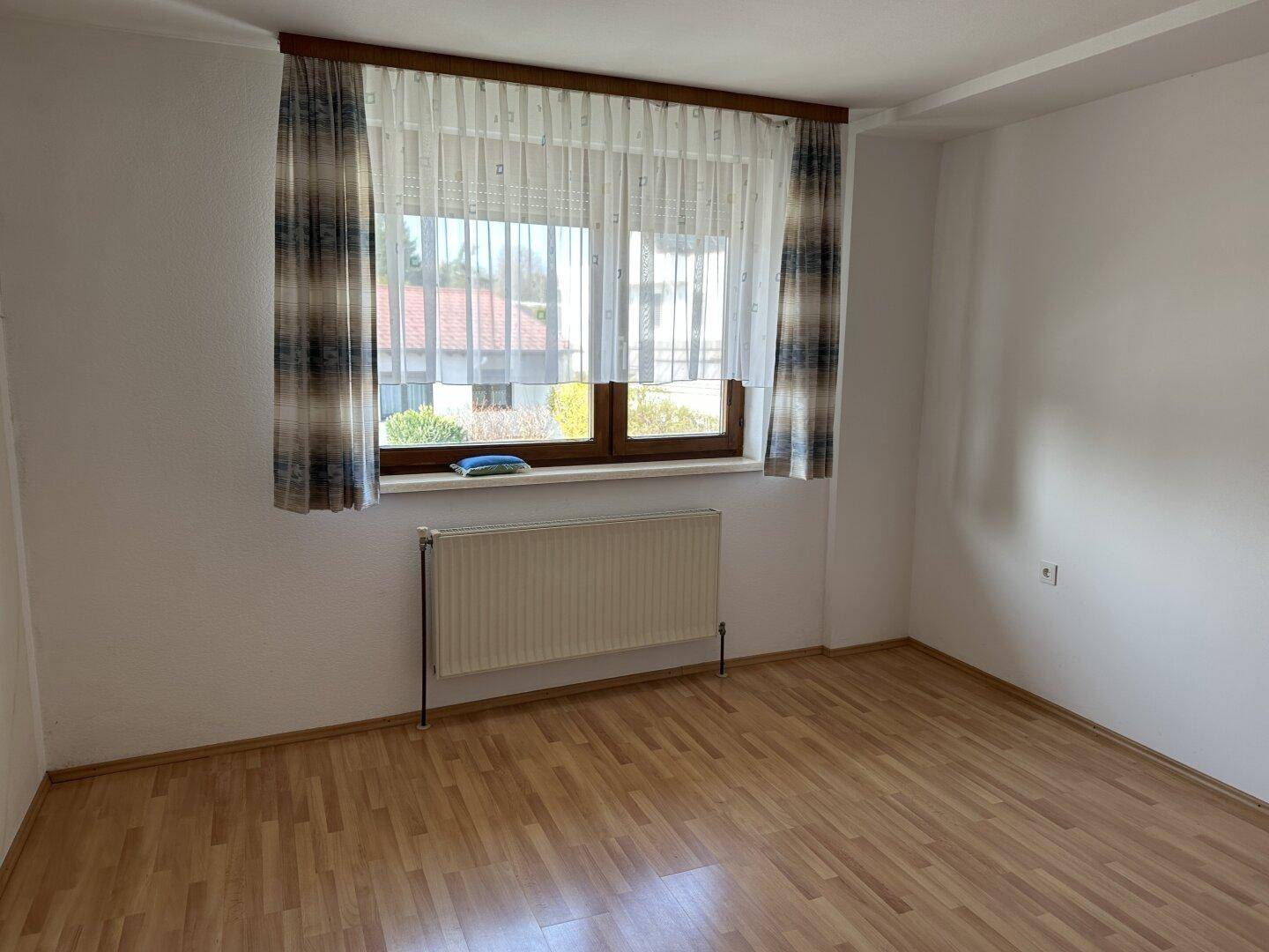 Leeres Zimmer mit Laminatboden, großem Fenster und Heizkörper.