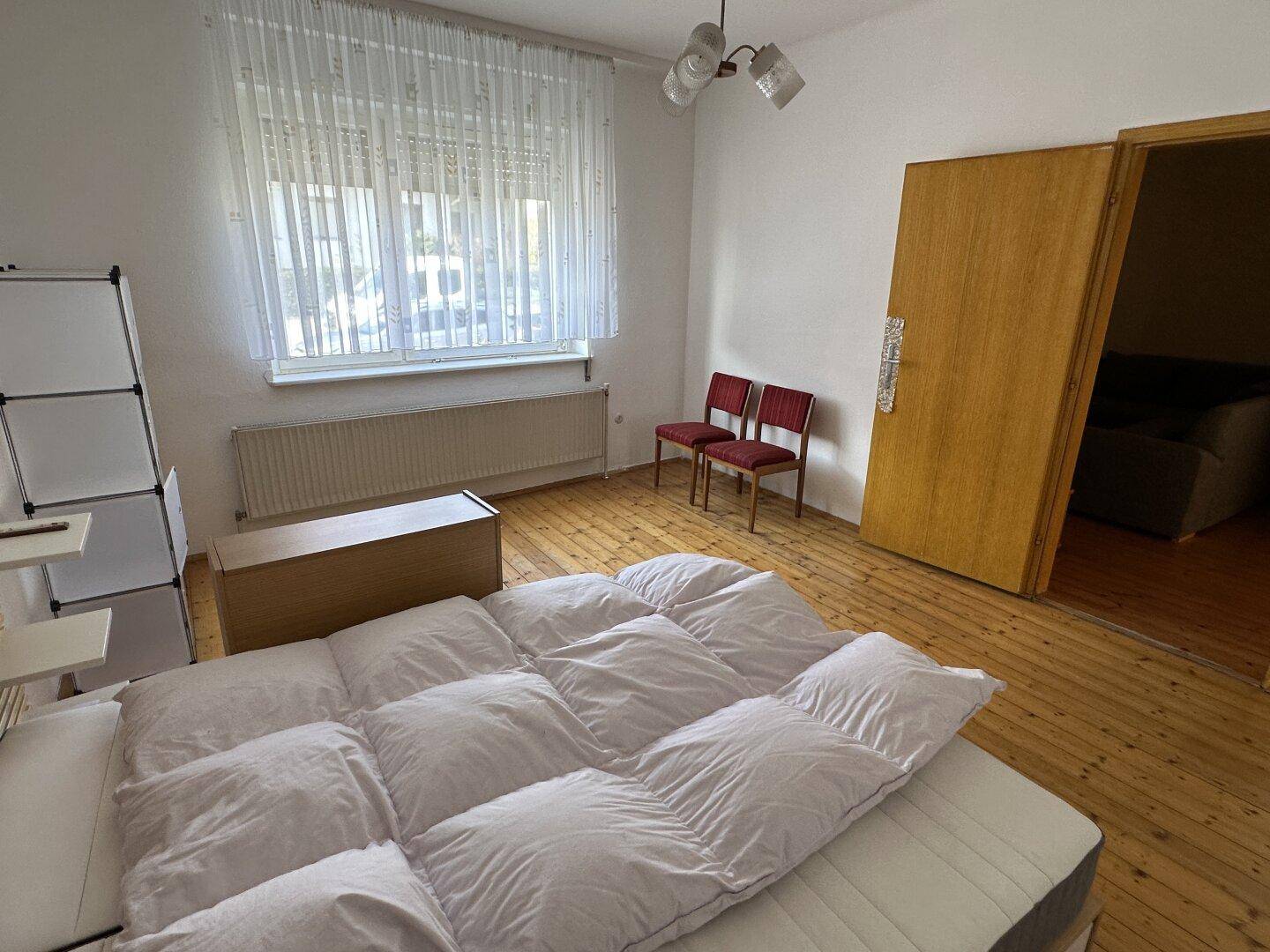 Schlafzimmer mit Holzboden, großem Fenster und einfacher Einrichtung.