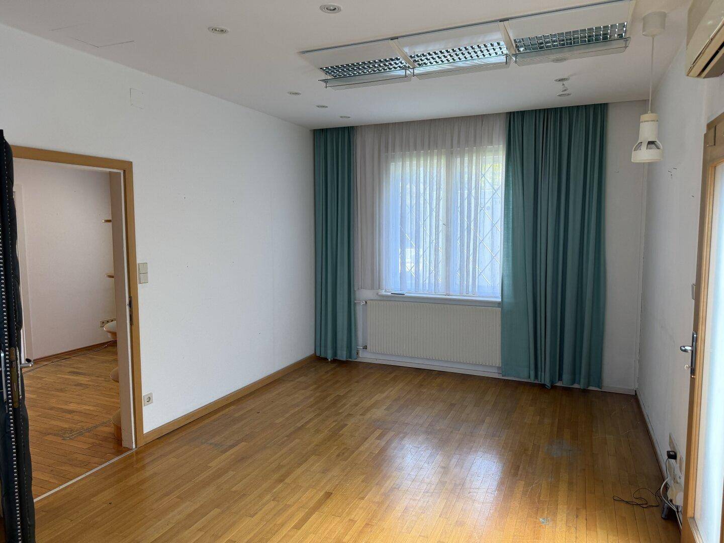 Heller Raum mit Holzparkettboden, großen Fenstern und Vorhängen, ideal als Büro oder Wohnzimmer.