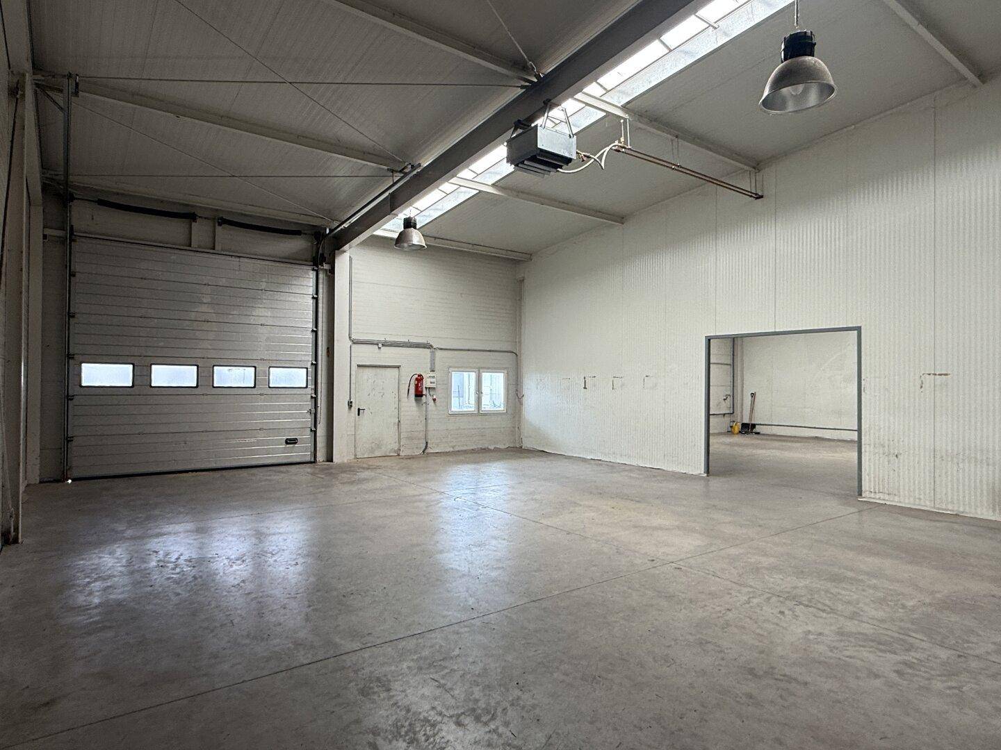 Blick in die Lagerhalle mit großem Rolltor, einem kleinen Fenster und einem offenen Zugang zu einem Nebenraum.
