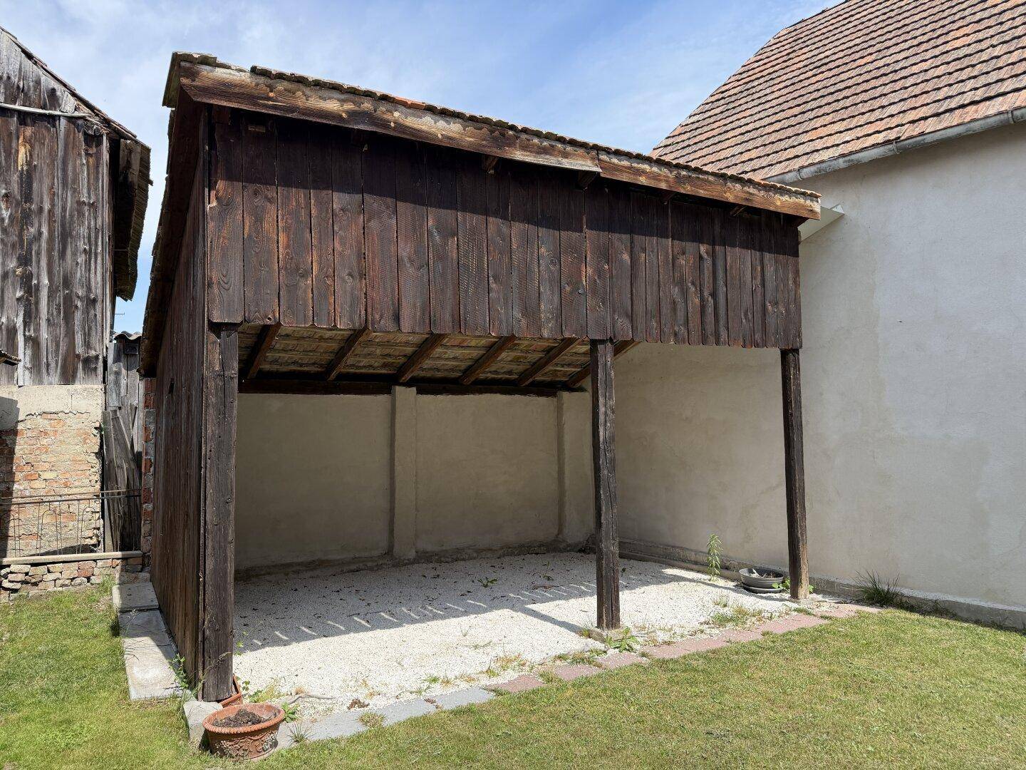 Offener Holzschuppen oder Carport mit Holzdach und Kiesboden im Gartenbereich.