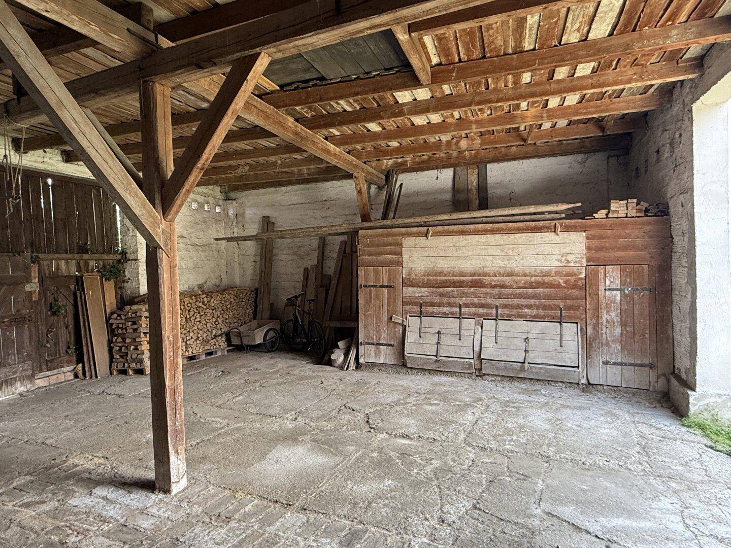 Geräumiger Innenraum eines Nebengebäudes mit Holzbalken, Betonboden und gelagertem Brennholz.