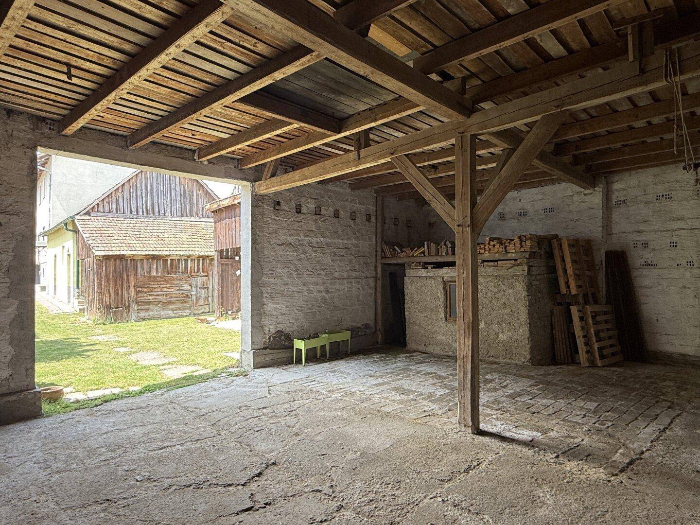 Innenansicht eines Nebengebäudes mit Holzbalken und Blick auf einen weiteren Holzschuppen im Freien.