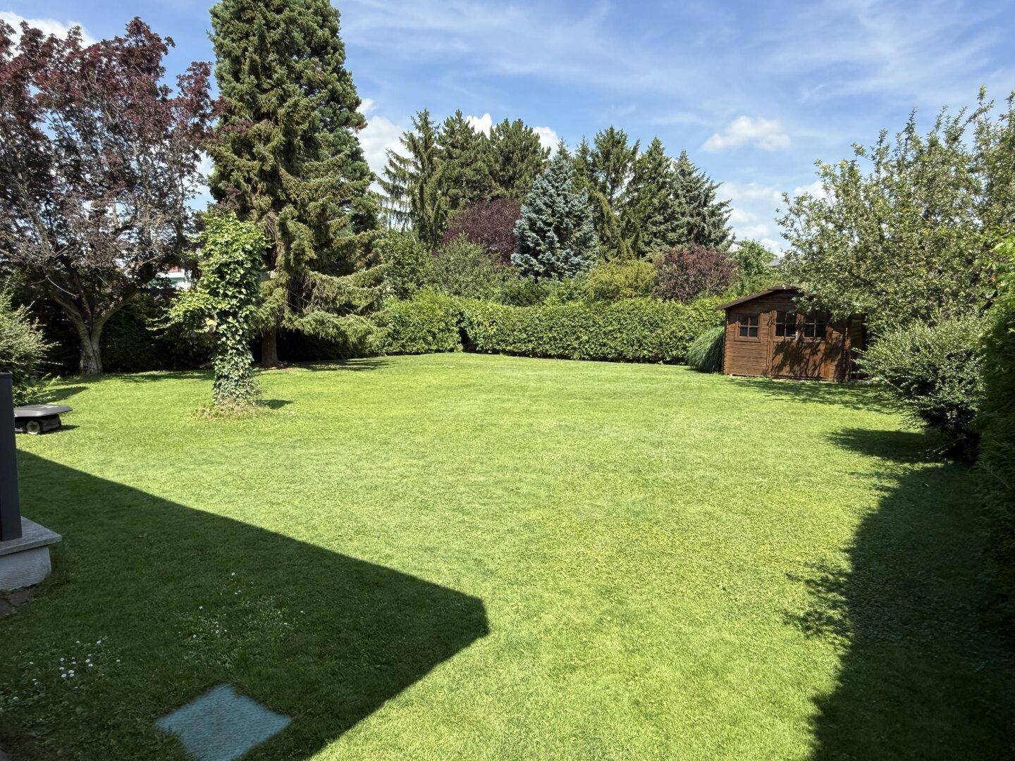 Weitläufiger Garten mit üppigem Rasen, hohen Bäumen und einem kleinen Gartenhaus.